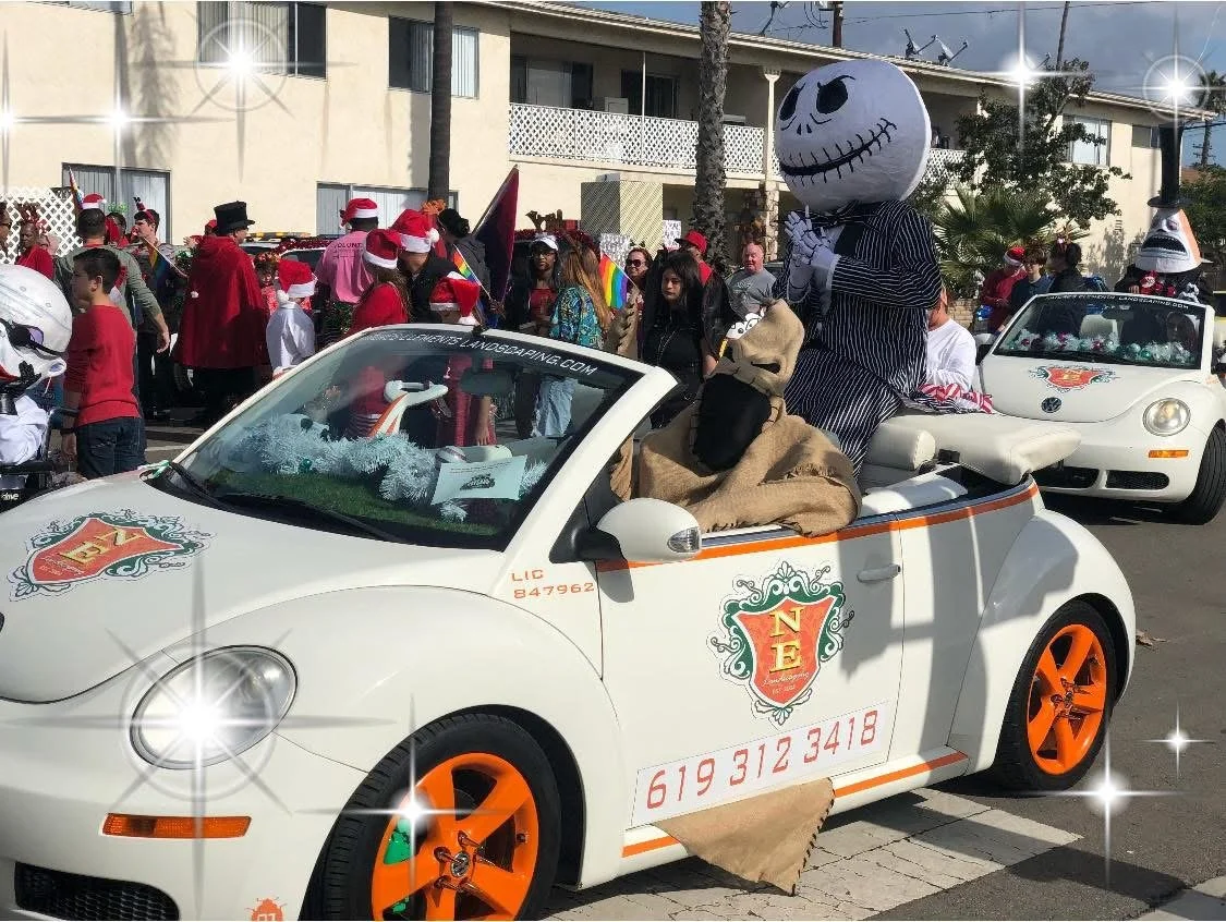 A parade with people dressed in holiday costumes, including Santa hats. There are decorated cars, one of which features a large skeleton mascot and a dog-like figure. Many spectators are along the street, some wearing Nightmare Before Christmas parad