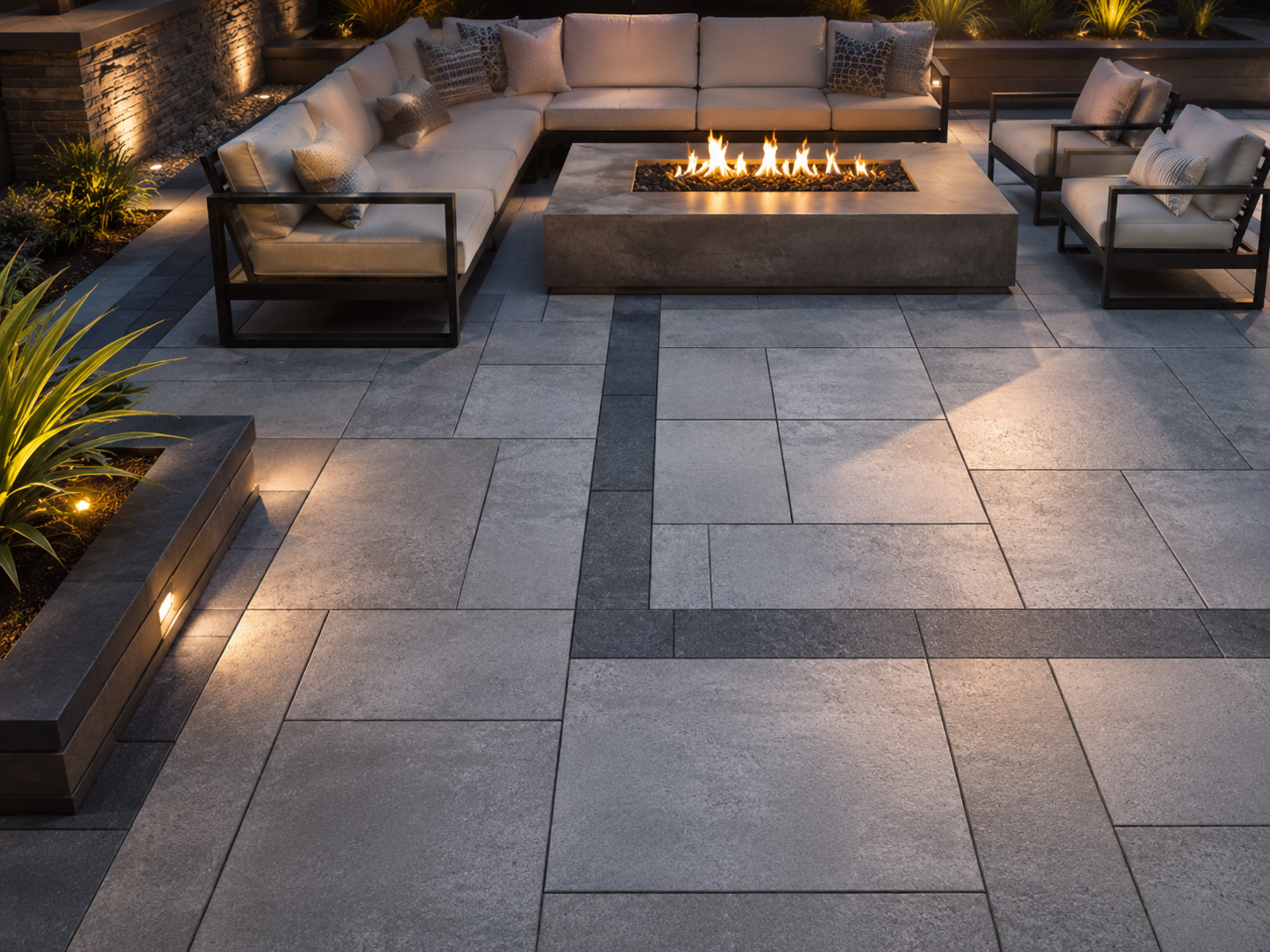 Outdoor paver patio with large square gray stone tiles, a rectangular fire pit in the center, and beige seating with throw pillows surrounding it. Plants and lighting add to the ambiance.
