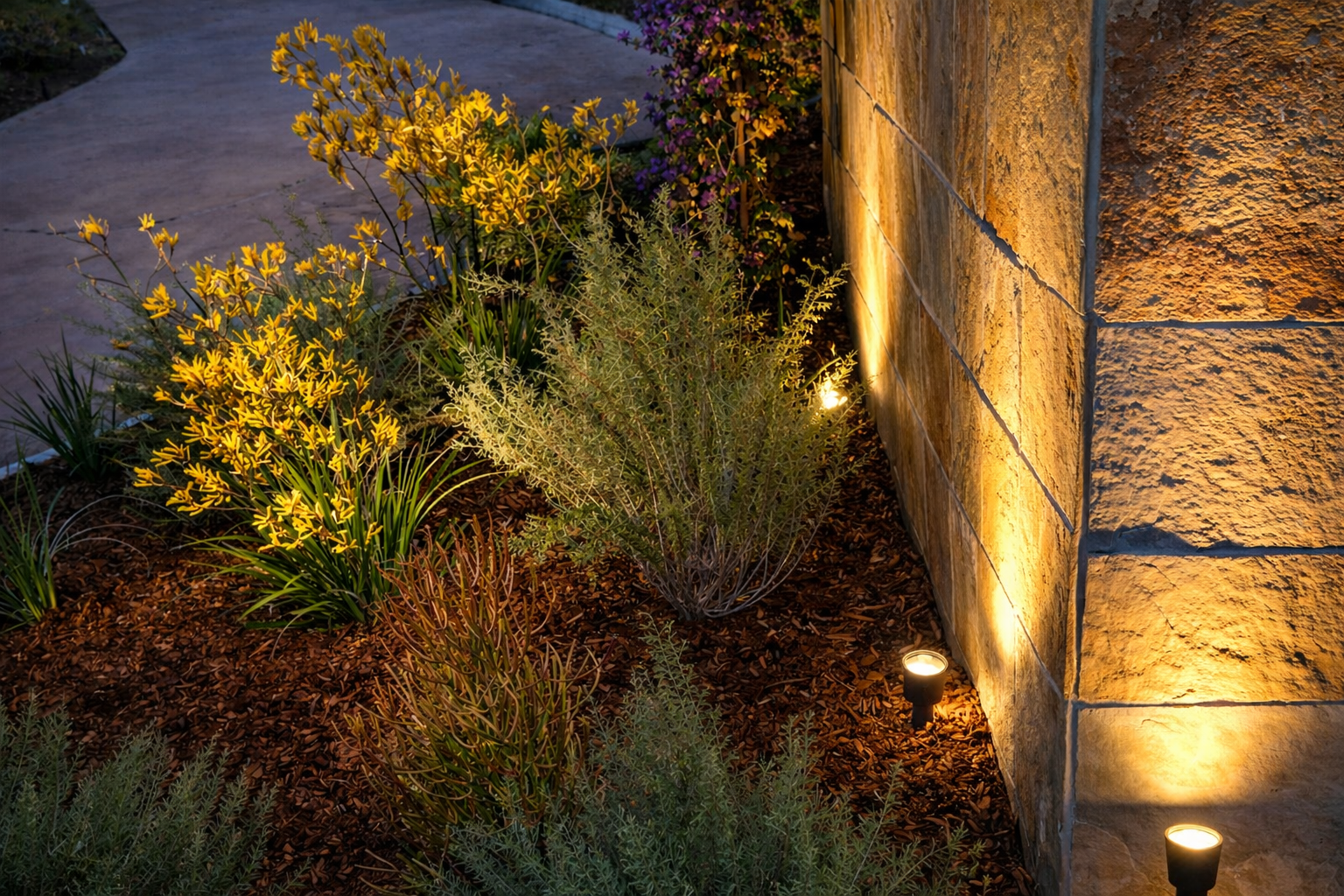 Outdoor garden with flowering plants illuminated by pathway lights next to a stone wall at dusk.