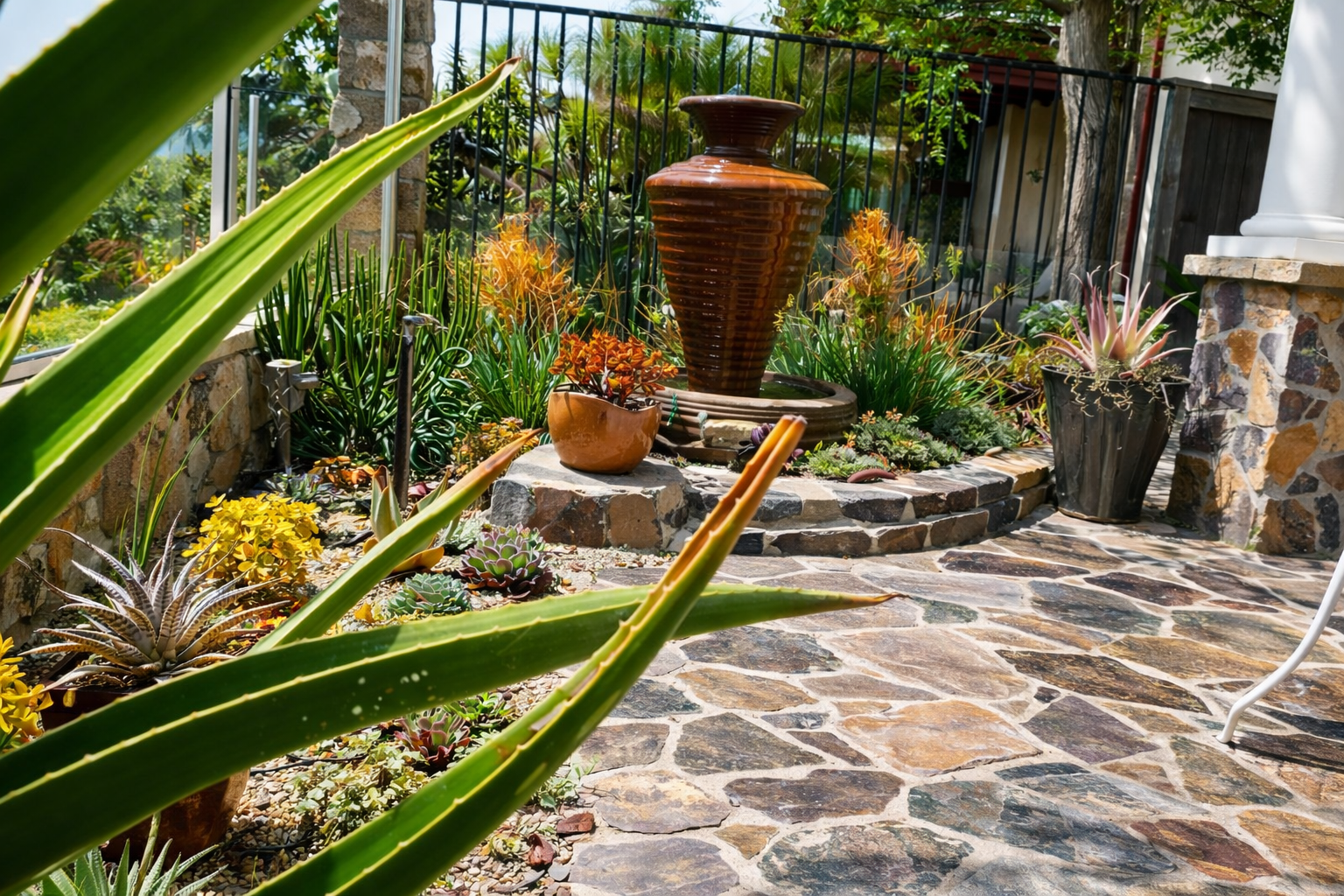A garden patio with drought-tolerant plants, succulents, and a central water fountain feature surrounded by stones, with a black metal fence and lush greenery in the background.