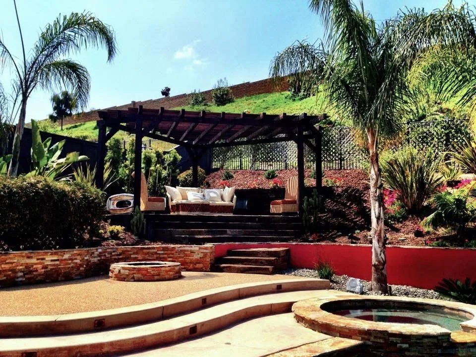 A backyard patio with a pergola-covered seating area on raised ground, surrounded by lush green plants and trees, with a multilevel, curved stone and brick landscape design, including a small round water feature in the foreground.  Pool and spa Contr