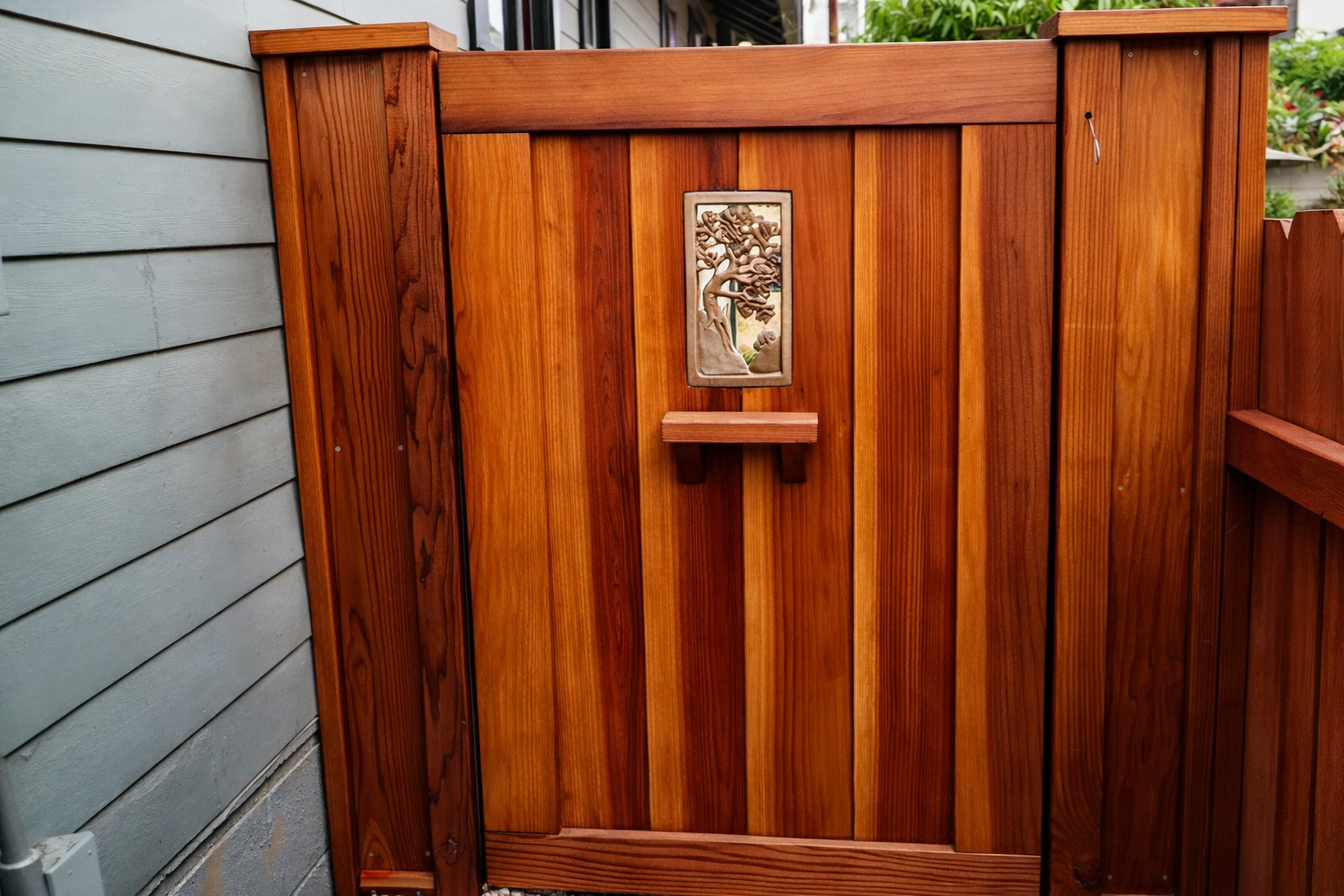 A wooden gate with a decorative plaque of a tree, attached to a gray house and fence, surrounded by greenery.
