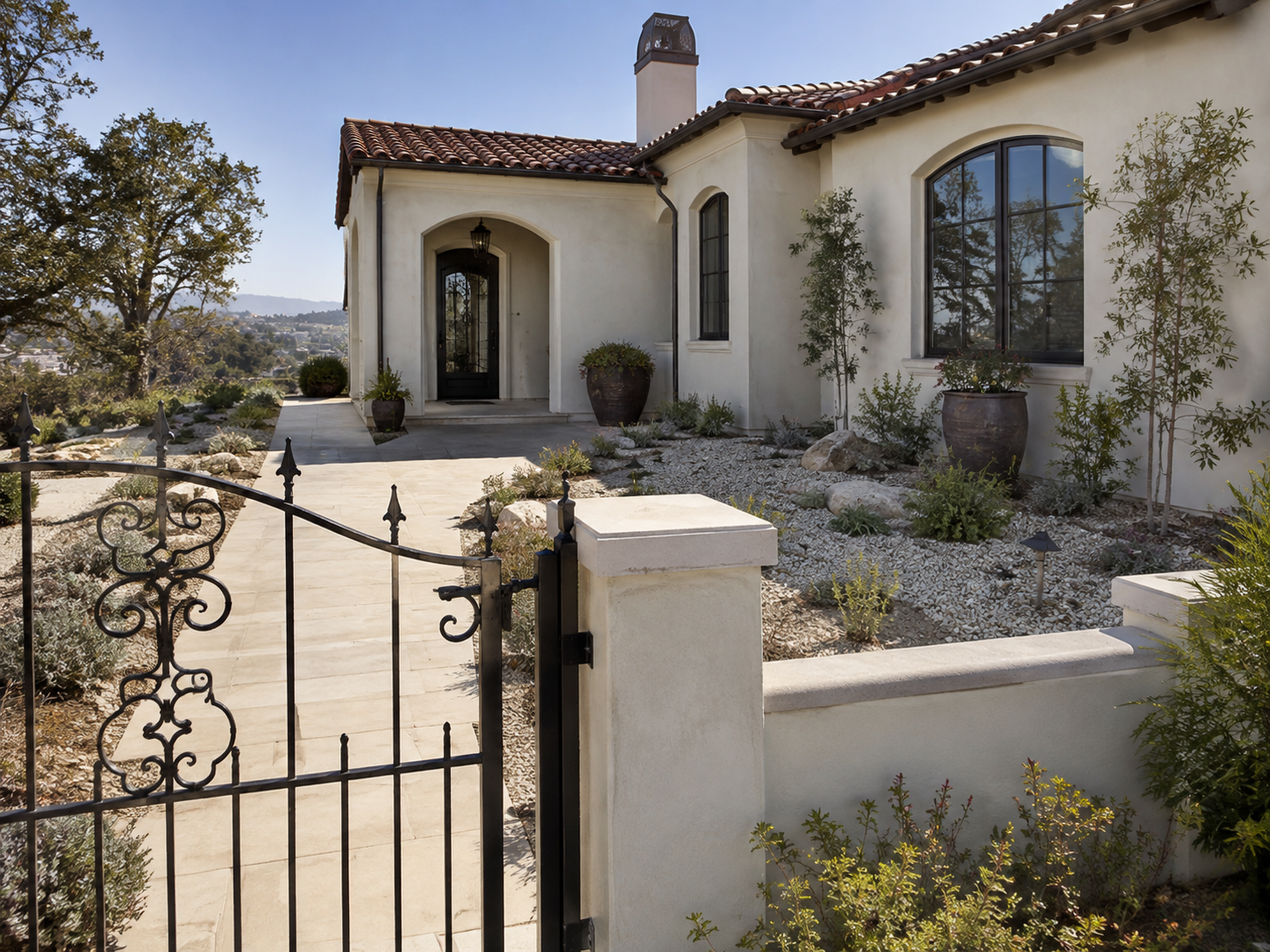 San Diego Zone 0 fire-conscious landscape design featuring a white stucco home with a red tile roof, black front door, and decorative metal gate surrounded by defensible space landscaping, drought-tolerant plants, gravel pathways, and low-maintenance
