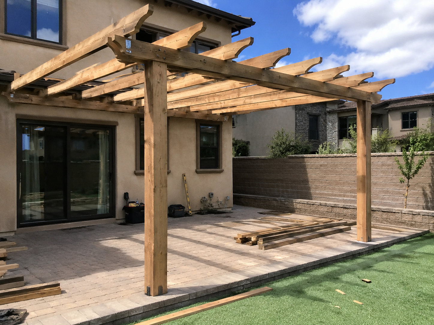 Construction of a wooden pergola on a patio of a house with brick flooring, neighboring houses in the background, and a blue sky with clouds.