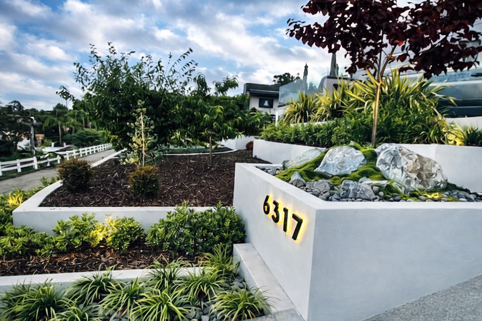 Modern landscaped garden with white concrete wall displaying house number 6317, filled with various plants, rocks, and trees on a cloudy sky day.  Contractor who can build a retaining wall near me. 