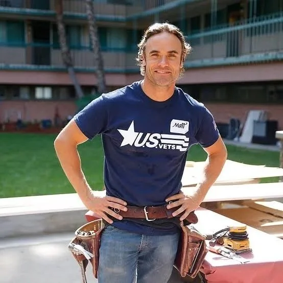 A man standing outdoors in front of a modern building, smiling, wearing a navy blue T-shirt with 'U.S. VETS' and a star logo, with a name tag that says 'Matt', and tools at his belt.