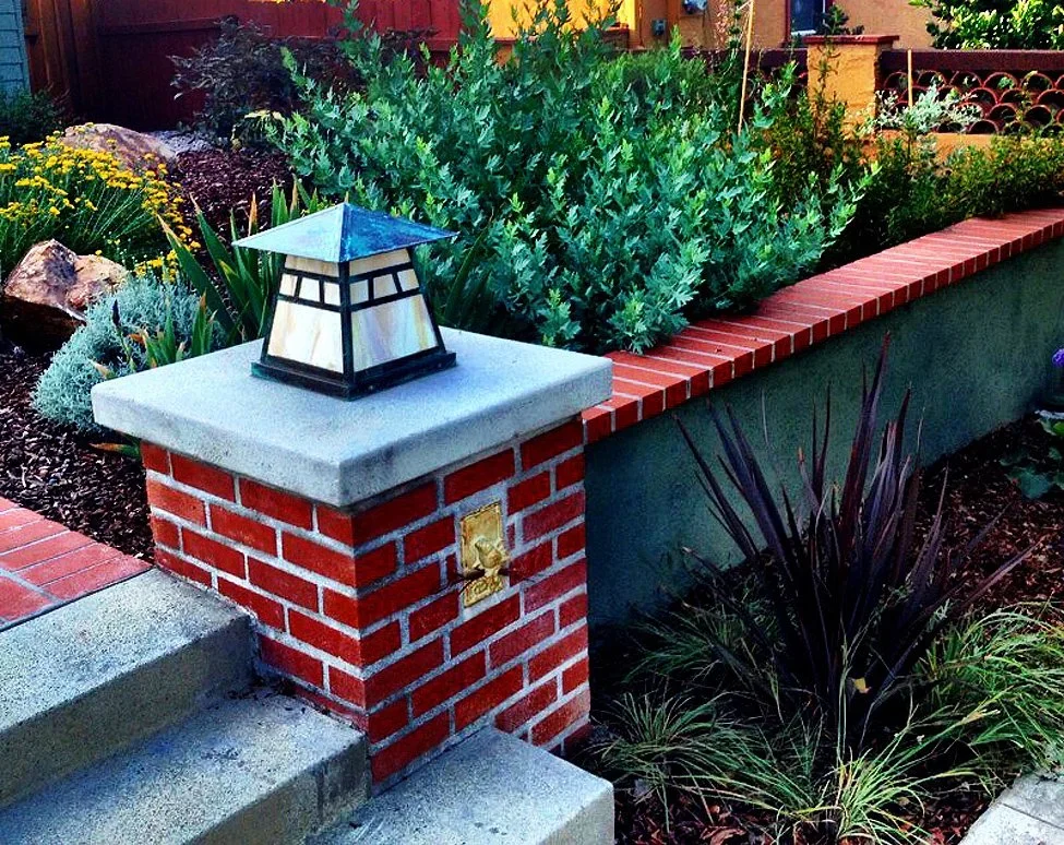 A landscaped front yard with a brick and concrete porch, a small turret-style lamp, and a raised garden bed filled with various green plants and shrubs.