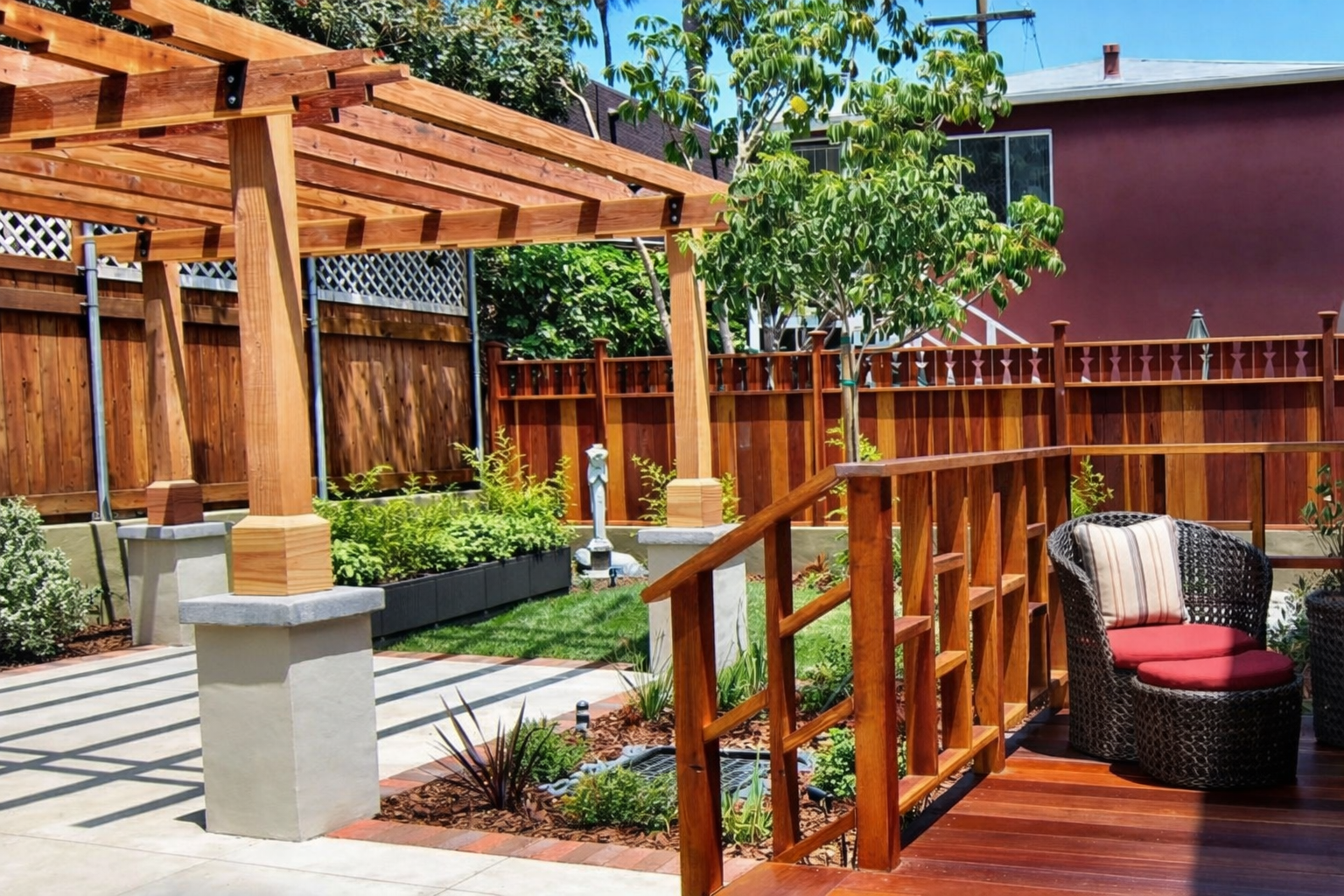 A backyard patio with a wooden deck, glass chair, and a garden area with plants, trees, and a wooden fence, under a clear blue sky.