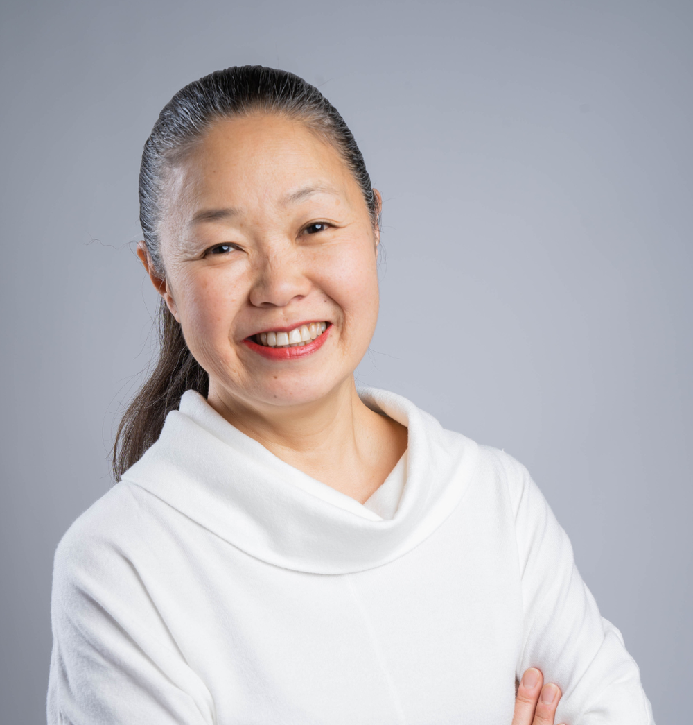 Makiko with dark hair pulled back, wearing a white top, standing against a grey background.