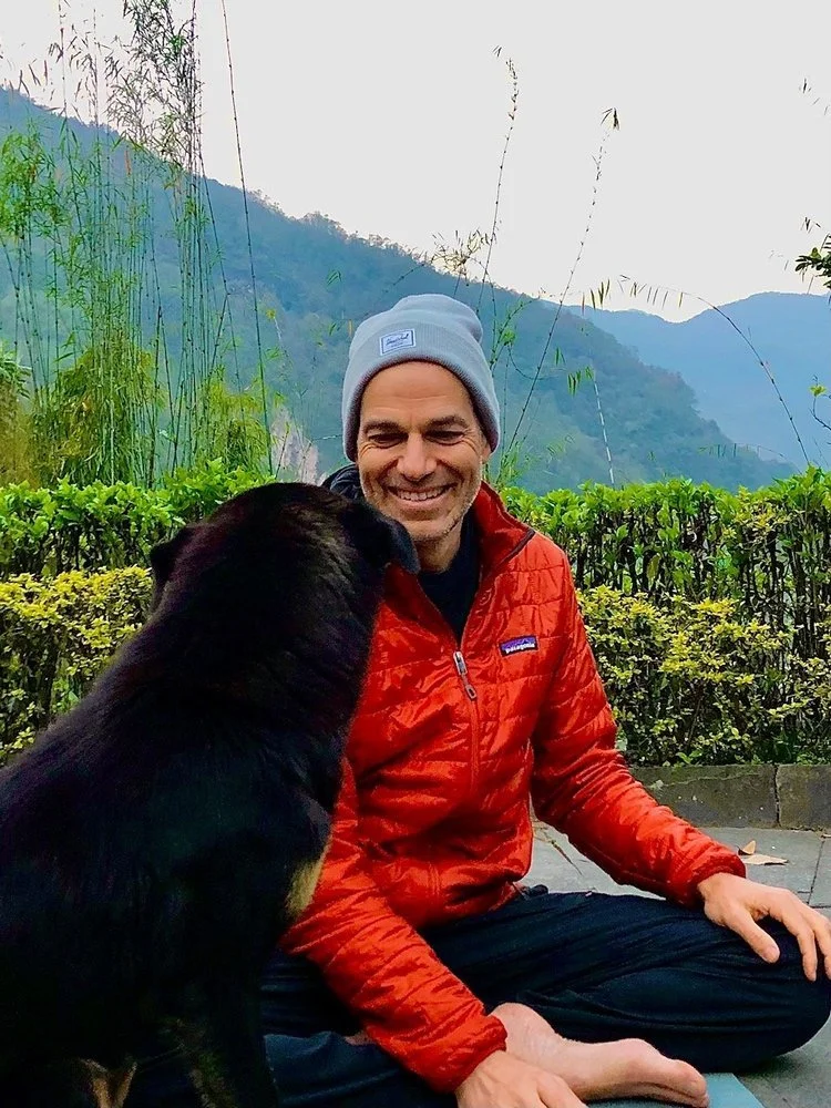 Rich Mancuso in a red jacket and gray beanie sitting outdoors on a stone surface, smiling, with a black dog sitting close beside him, surrounded by green bushes and mountains in the background.