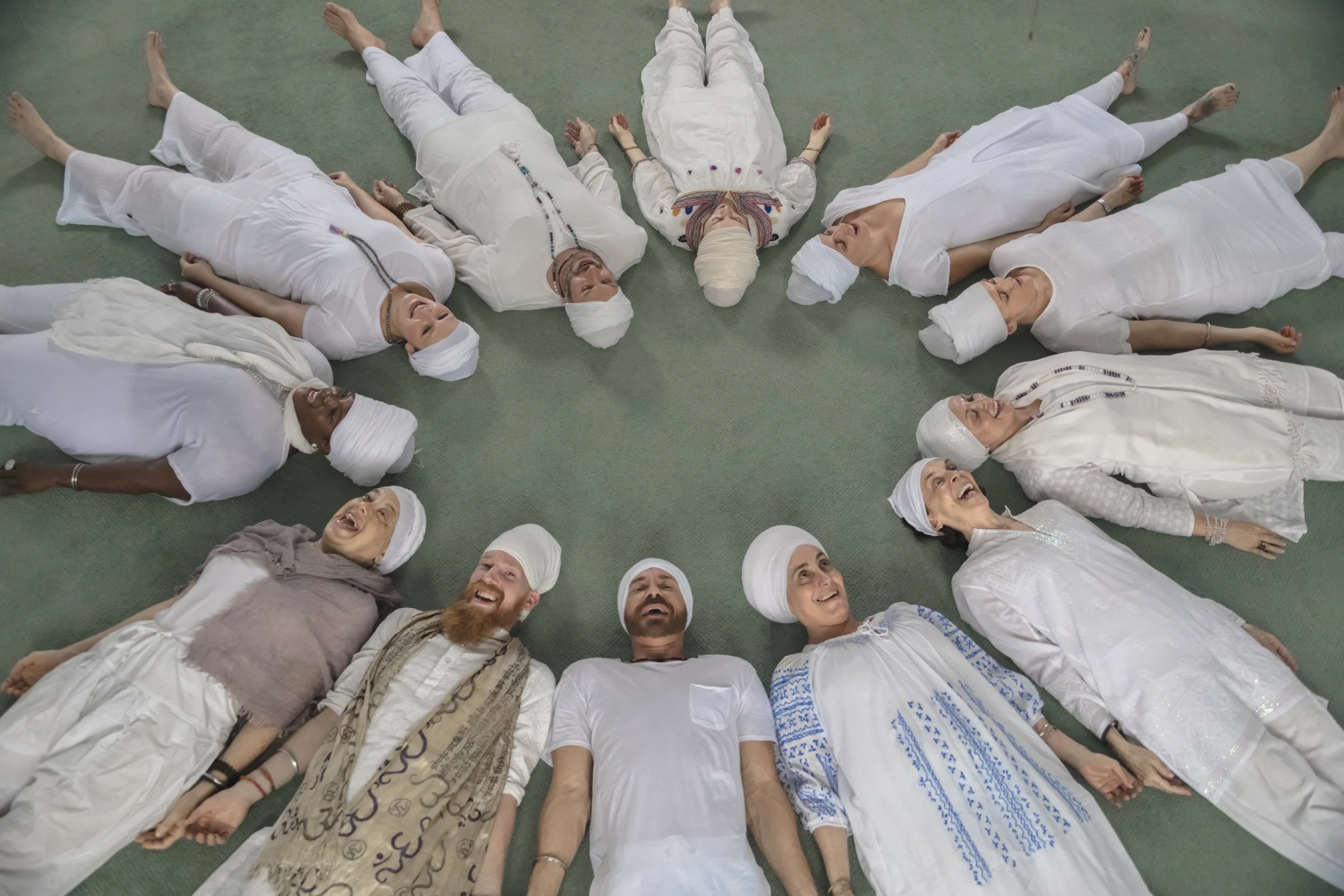 Group of diverse people lying on the floor in a circle, smiling and looking up, dressed in white traditional or spiritual clothing.