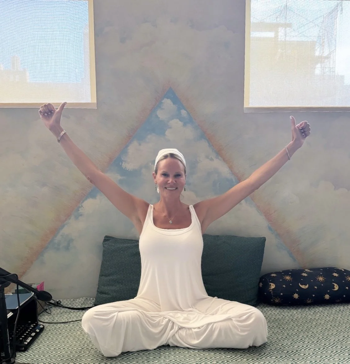 Jessica sitting cross-legged on a bed with arms raised in a victory pose, smiling, in front of a painted wall mural of a sky with clouds, with two windows above.