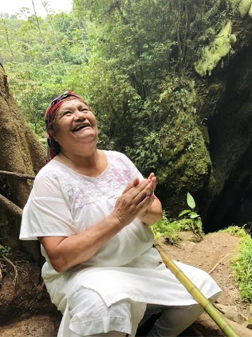 An elderly woman with a colorful headscarf, wearing a white dress, sitting outdoors in a lush, green forest, smiling joyfully with her hands clasped, surrounded by rocks and trees.