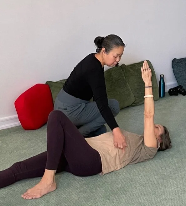 Two women practicing yoga on a green carpeted floor, one lying down with her arm raised, the other assisting in a stretch.