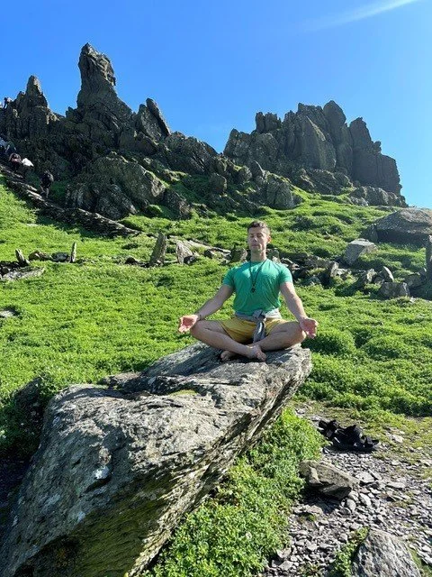 Person sitting cross-legged on a large rock in a green, mountainous landscape, practicing meditation under a bright blue sky with rocky peaks in the background.
