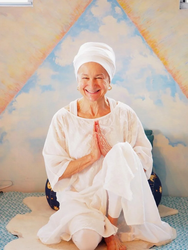 Gail/Atma Dharam Kaur wearing a white headwrap and white clothing, sitting on a patterned mat with hands in prayer pose, in front of a colorful wall mural.
