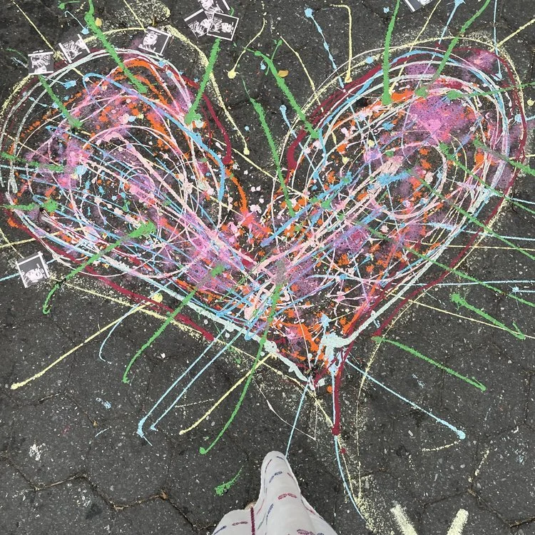 Colorful heart-shaped chalk drawing on pavement with paint splatters and spray paint marks, with a person's feet visible at the bottom.