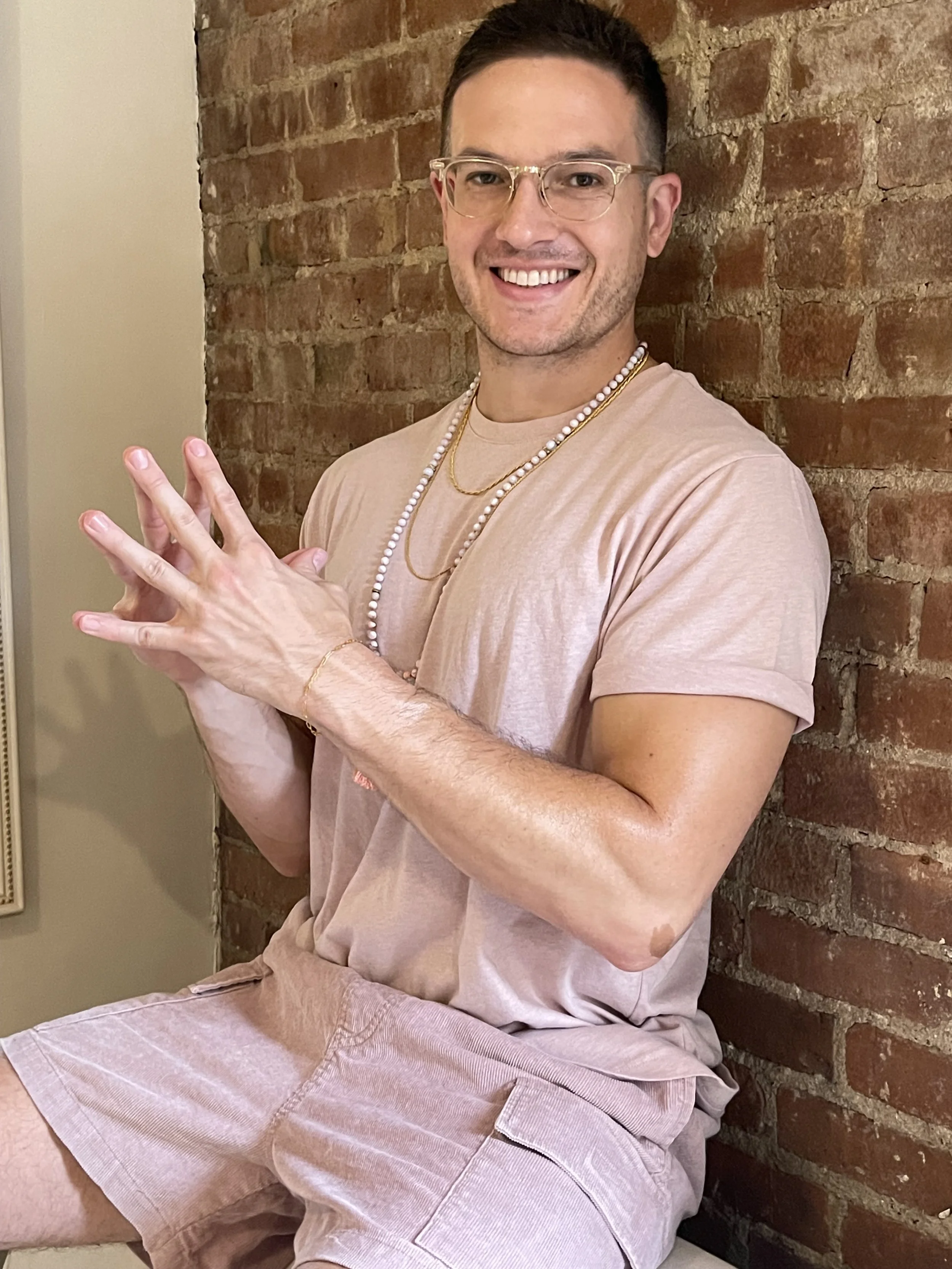 Josh/Tej Randeep with glasses, tattoos, and jewelry, wearing a pink t-shirt and shorts, sitting against a brick wall.