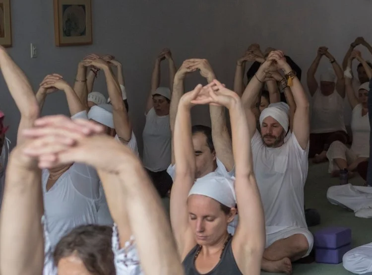 Group of people practicing yoga in a studio, seated with arms raised overhead and hands clasped, wearing white and black athletic clothing, some with head coverings.
