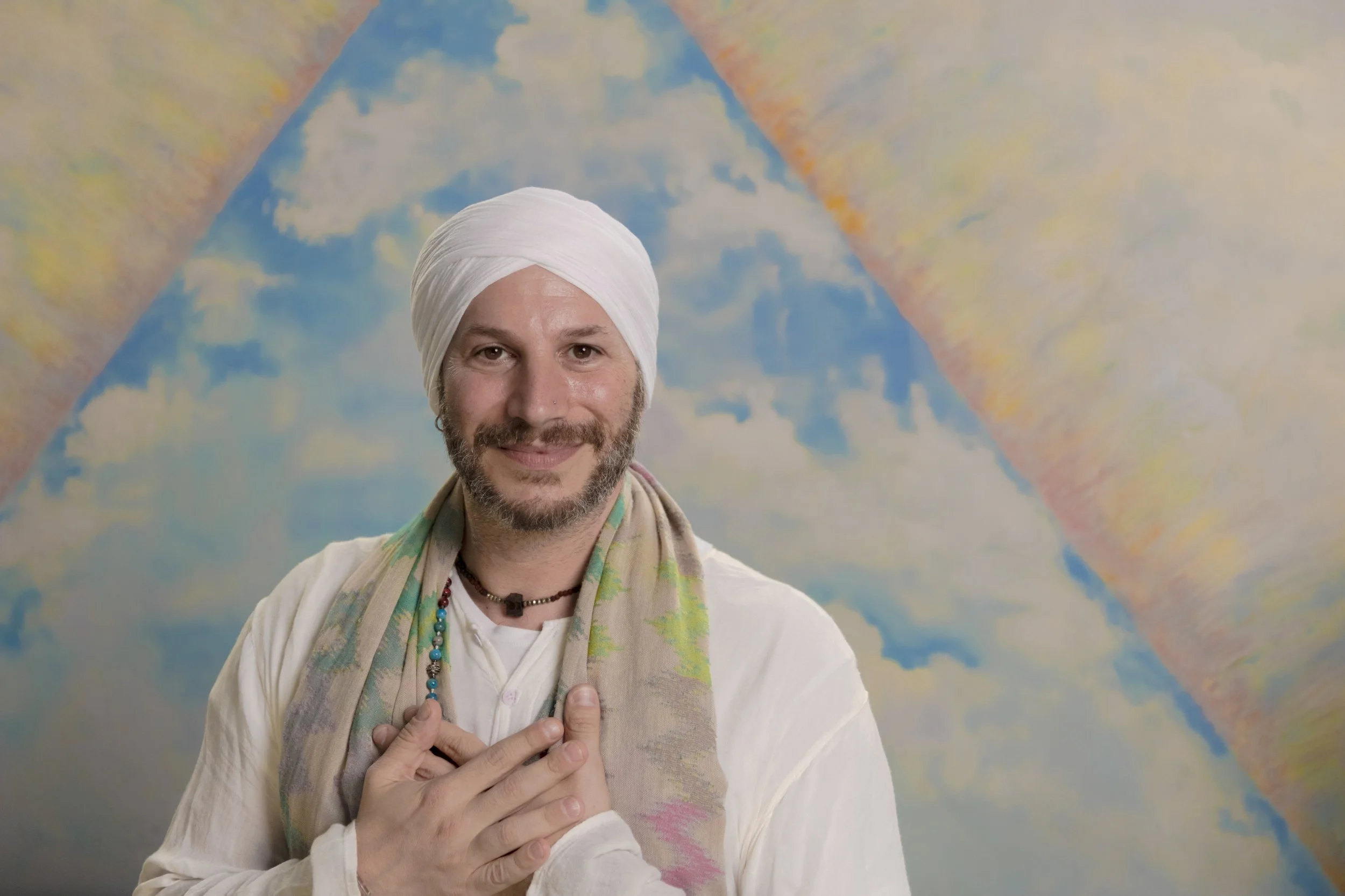Raj Karam wearing a white turban, a beard, and a friendly smile standing in front of a colorful cloud and sky mural background. He is wearing a cream-colored shirt, a multicolored scarf, and beaded necklaces, with his hands placed over his chest.