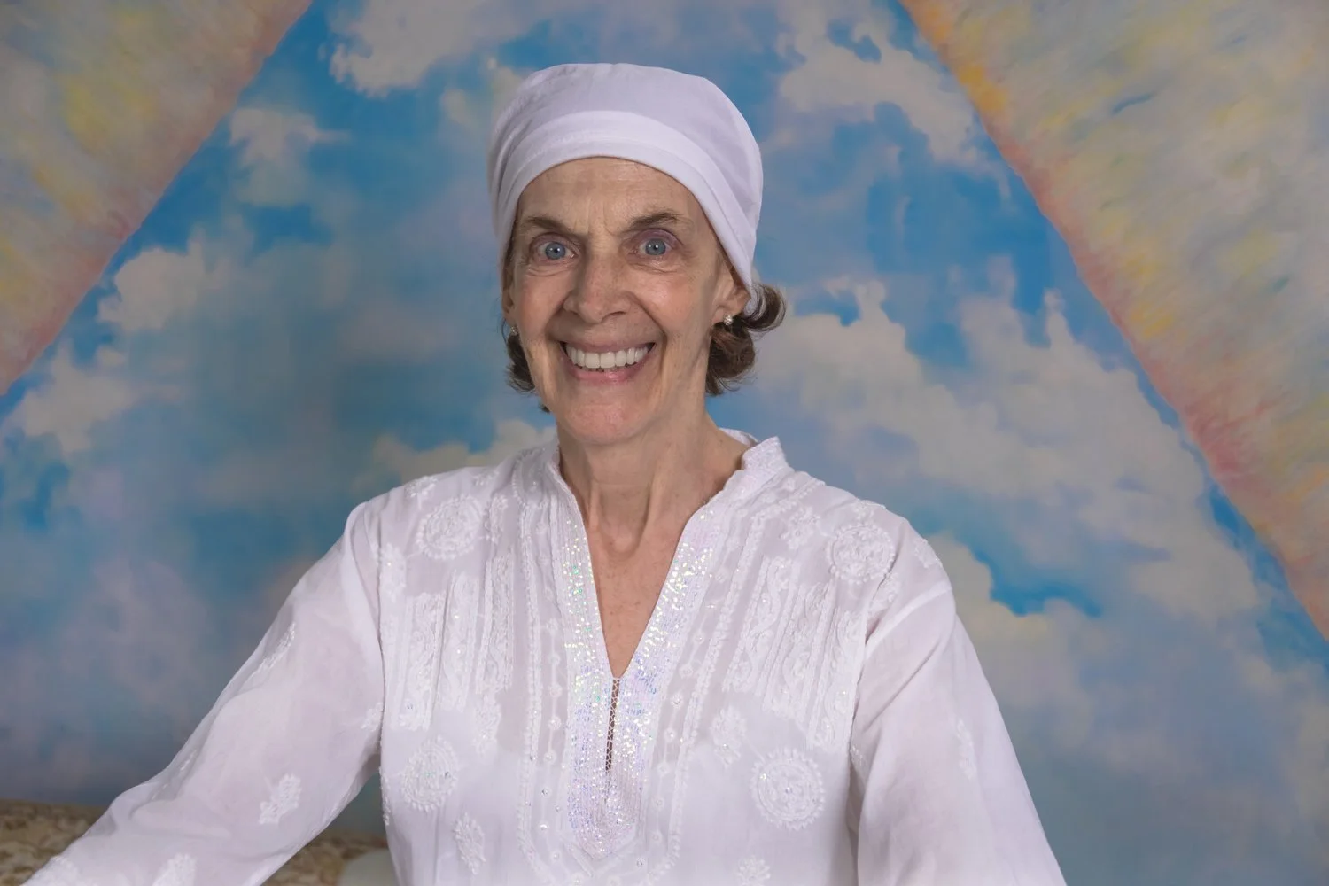 Deborah/Harinam wearing a white headscarf and white embroidered top, smiling in front of a colorful sky mural background.