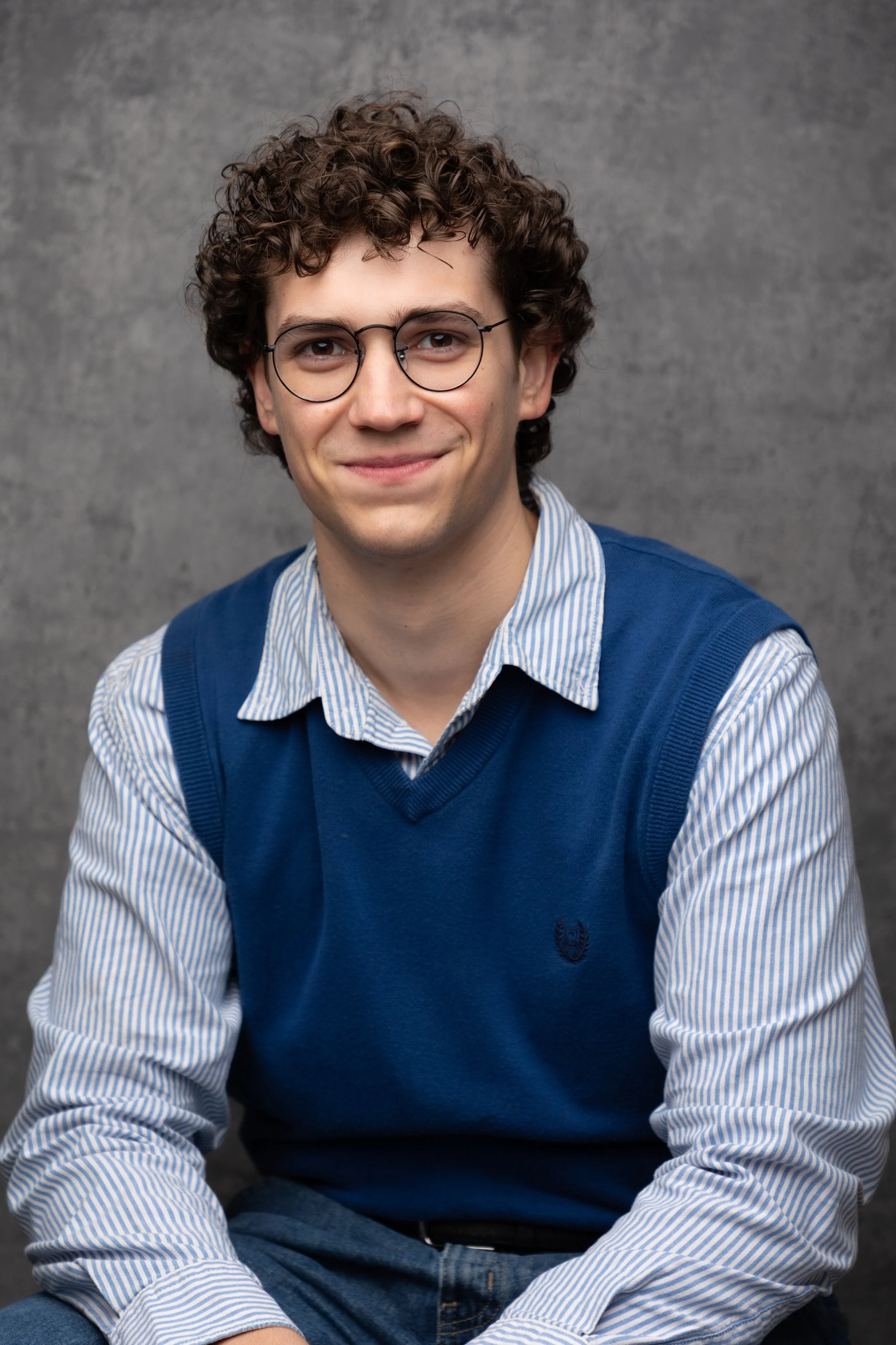 Young man with curly brown hair, round glasses, wearing a blue vest over a striped shirt, sitting against a gray background, smiling gently.