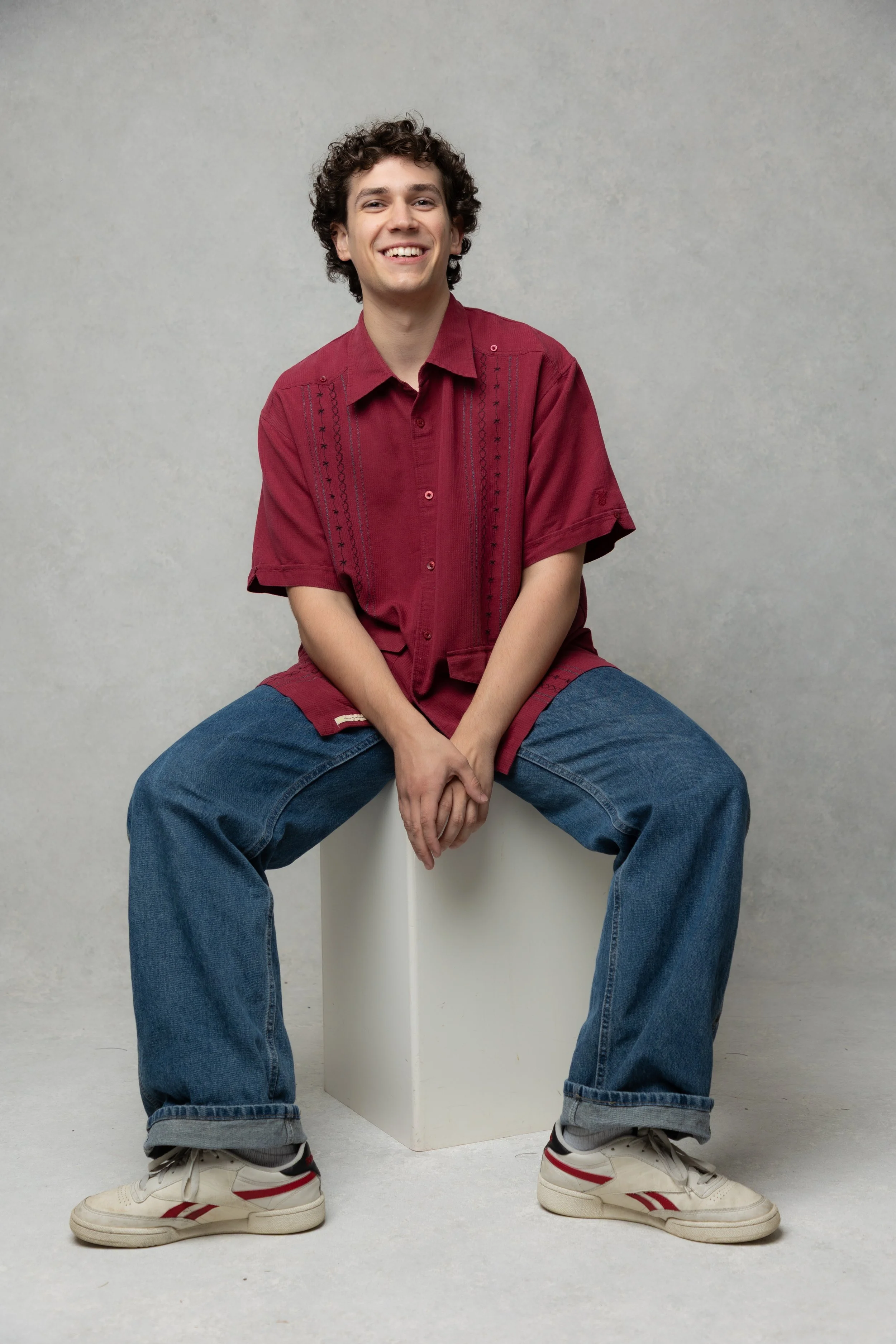 Young man with curly hair smiling, wearing a red shirt and jeans, sitting on a white platform against a plain gray background.