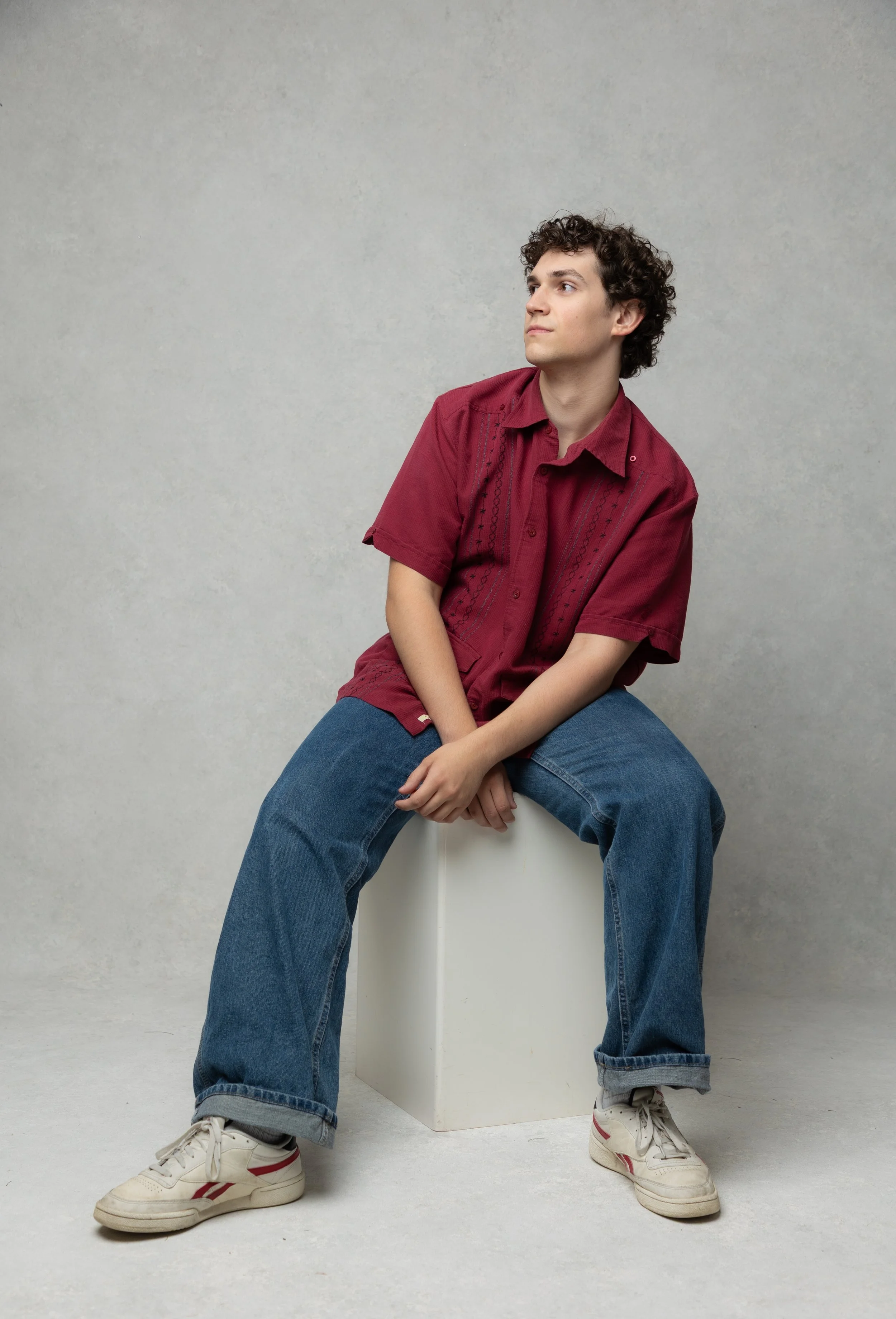 A young man with curly dark hair sitting on a white cube, wearing a red short-sleeved shirt with embroidery, blue jeans, and white sneakers with red and gray accents, against a gray textured background.