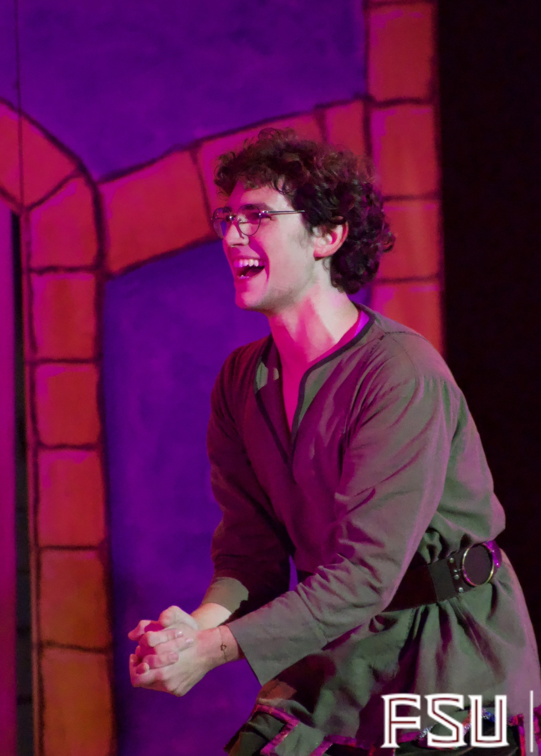 A young man with curly dark hair and glasses, wearing an olive green dress with a black belt, is smiling and appears to be in a theatrical performance, standing in front of a colorful painted backdrop with red bricks and purple and blue tones.