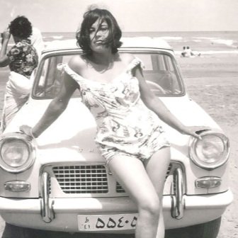 A woman in a vintage swimsuit leans against the front of a classic car on a beach, with another person and the ocean in the background.