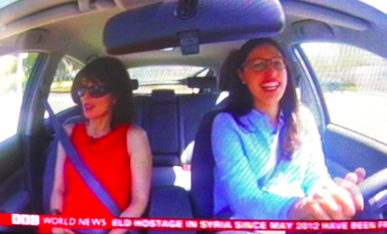 Two women sitting in a car, both wearing seatbelts; one is driving and smiling, the other is in the passenger seat wearing sunglasses. A news ticker is visible at the bottom of the image.