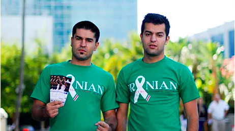 Two men wearing green T-shirts, holding papers, standing outdoors.