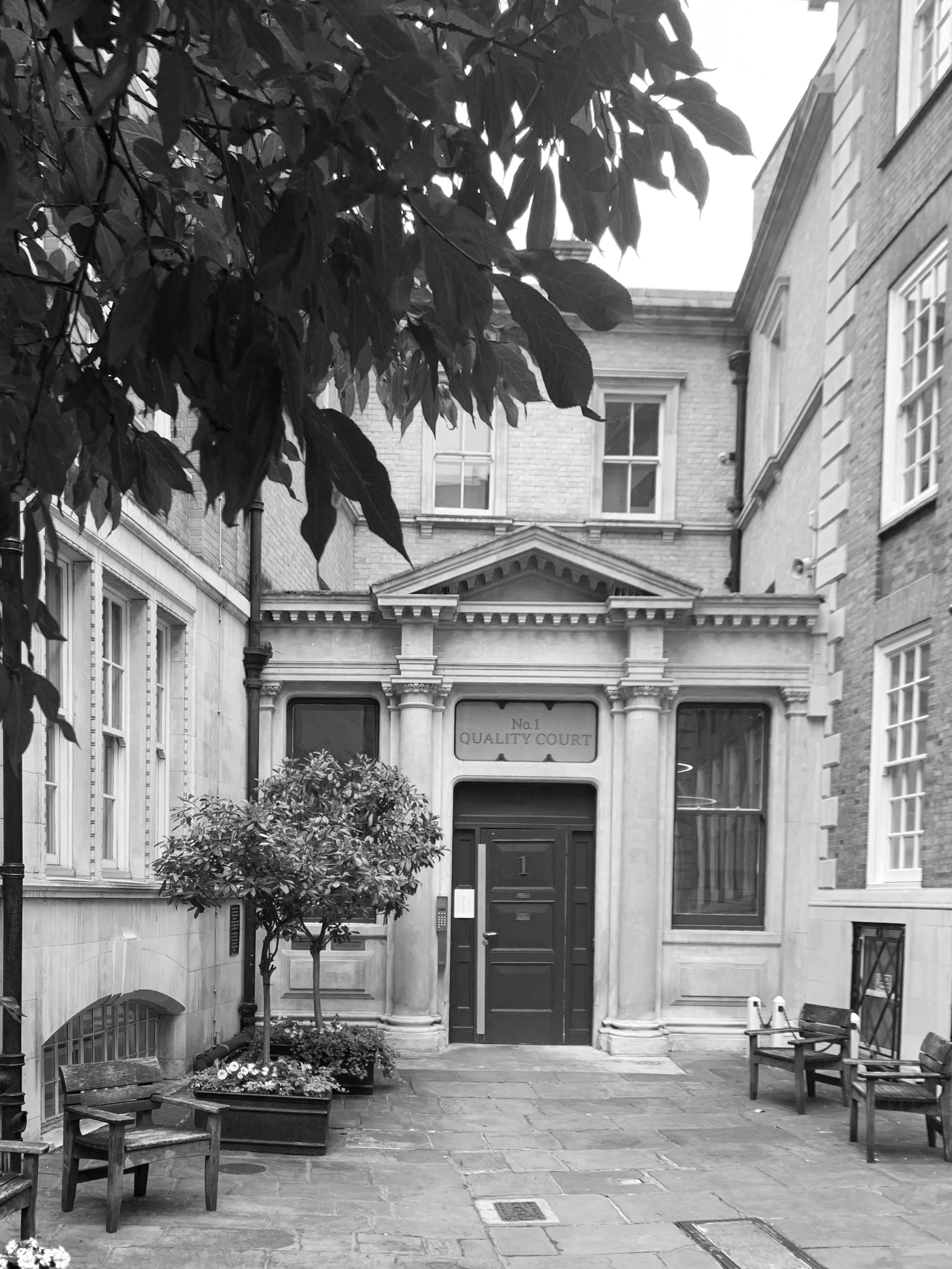 Black and white photo of the entrance to a building labeled 'No.1 Quality Court,' with benches, potted plants, and trees in a courtyard.