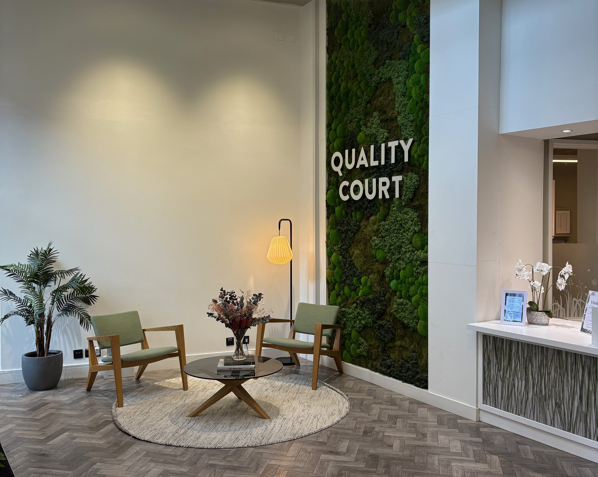 Lobby area with two green armchairs, a round coffee table with a book and a flower arrangement, a tall green potted plant, and a moss wall with the words 'Quality Court' on it.