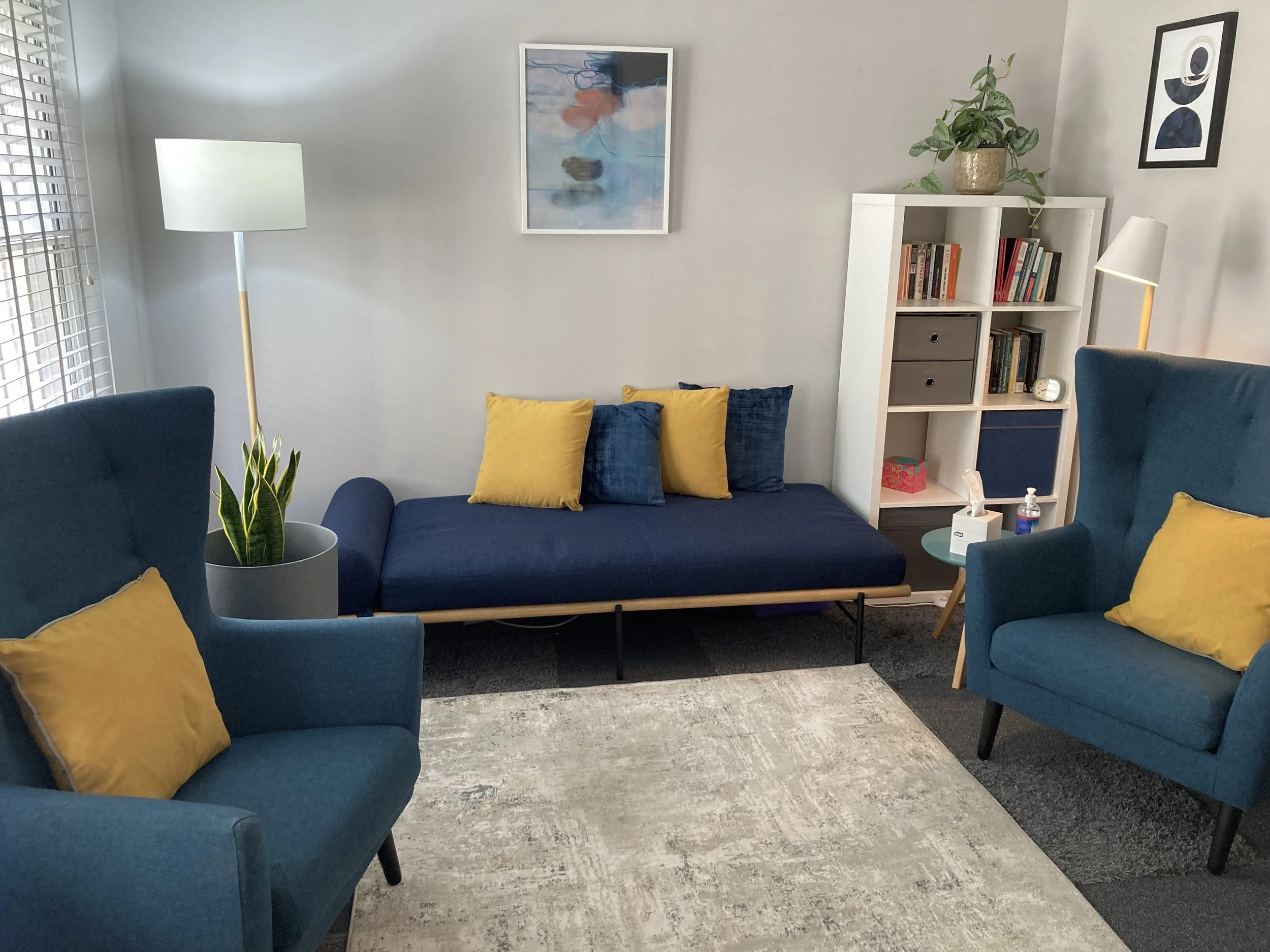 Living room with two blue armchairs and one blue daybed with yellow and blue pillows. There are two floor lamps, a white bookshelf with books and decor, a potted plant, and artwork on light gray walls. A window with white blinds is on the left.