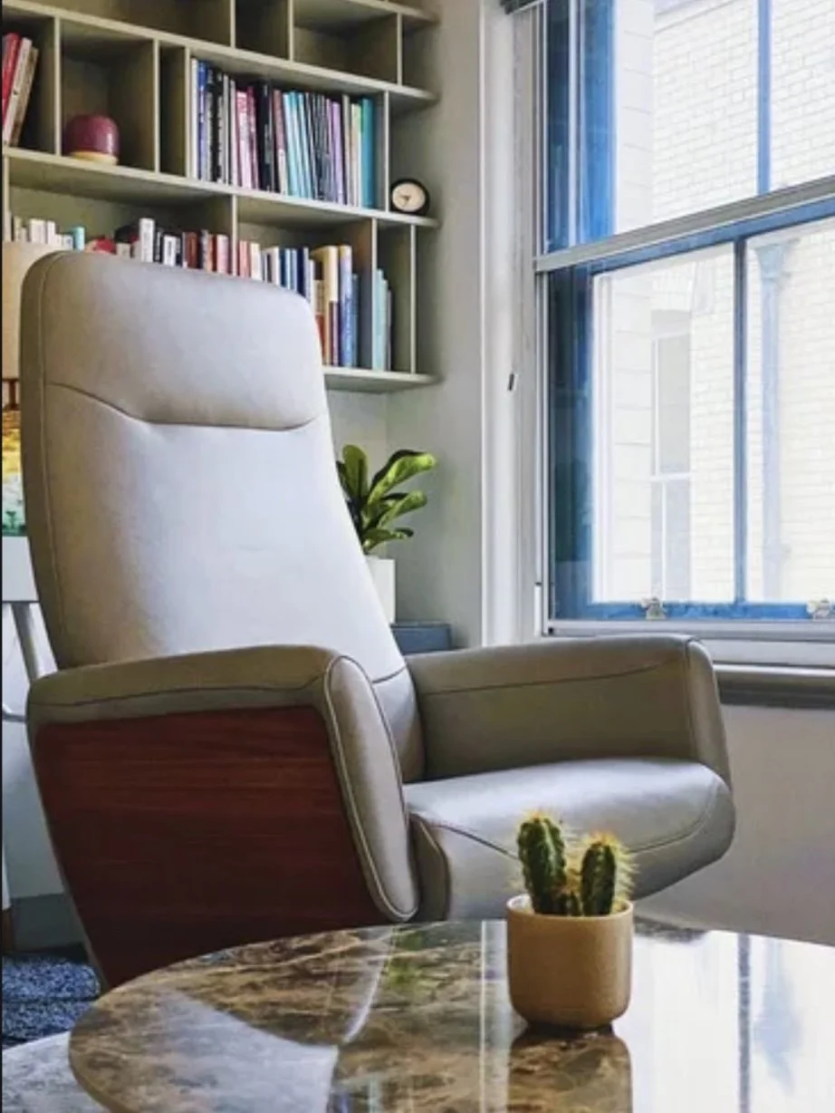 A cozy reading area featuring a beige recliner chair near a window with shelves of books above. A small cactus plant is on a marble table in the foreground.
