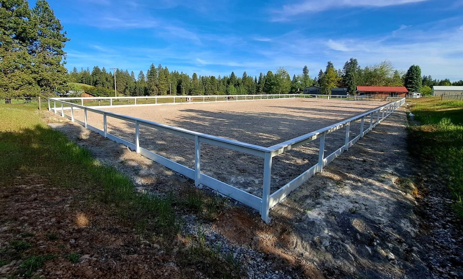 Empty outdoor arena with a white railing, surrounded by green trees, farm buildings, and a blue sky.