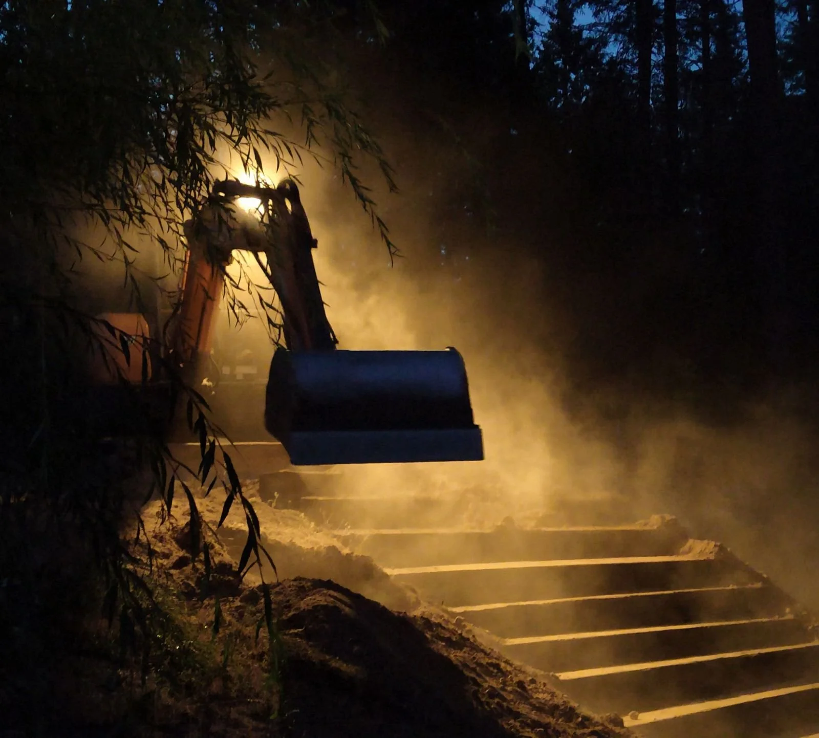 Backhoe digging into the ground at night, with dust and dirt being illuminated by a bright light, surrounded by trees.