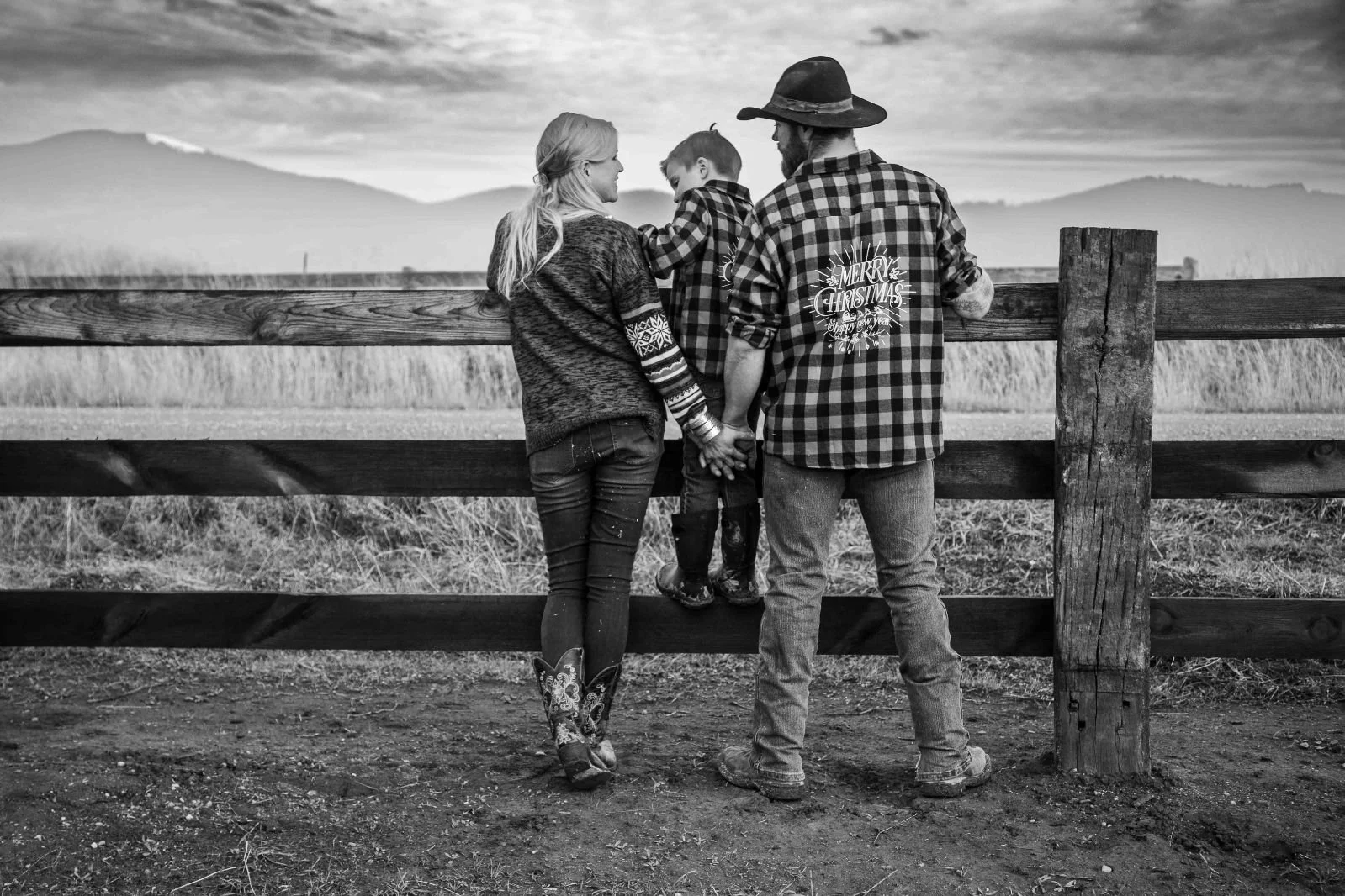 A family of three, a woman, a man, and a child, standing behind a wooden fence in a rural field during dusk. The woman has long hair and is wearing a patterned sweater and jeans. The man is wearing a hat and a checkered shirt with a birthday message on the back. The child, who is in the middle, is leaning against the woman and is being held by the man. They are holding hands, and the landscape shows rolling hills in the background.