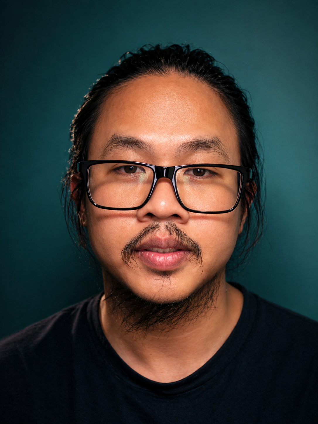 Close-up portrait of a man with glasses, facial hair, and long black hair against a dark teal background.