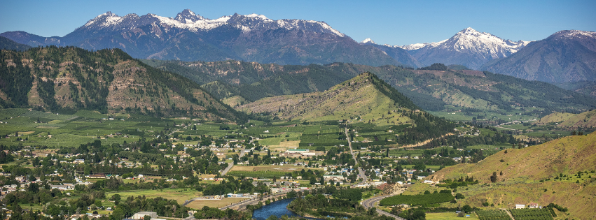 Scenic view of a valley surrounded by mountains with snow-capped peaks, green fields, and a small town with a river running through it.