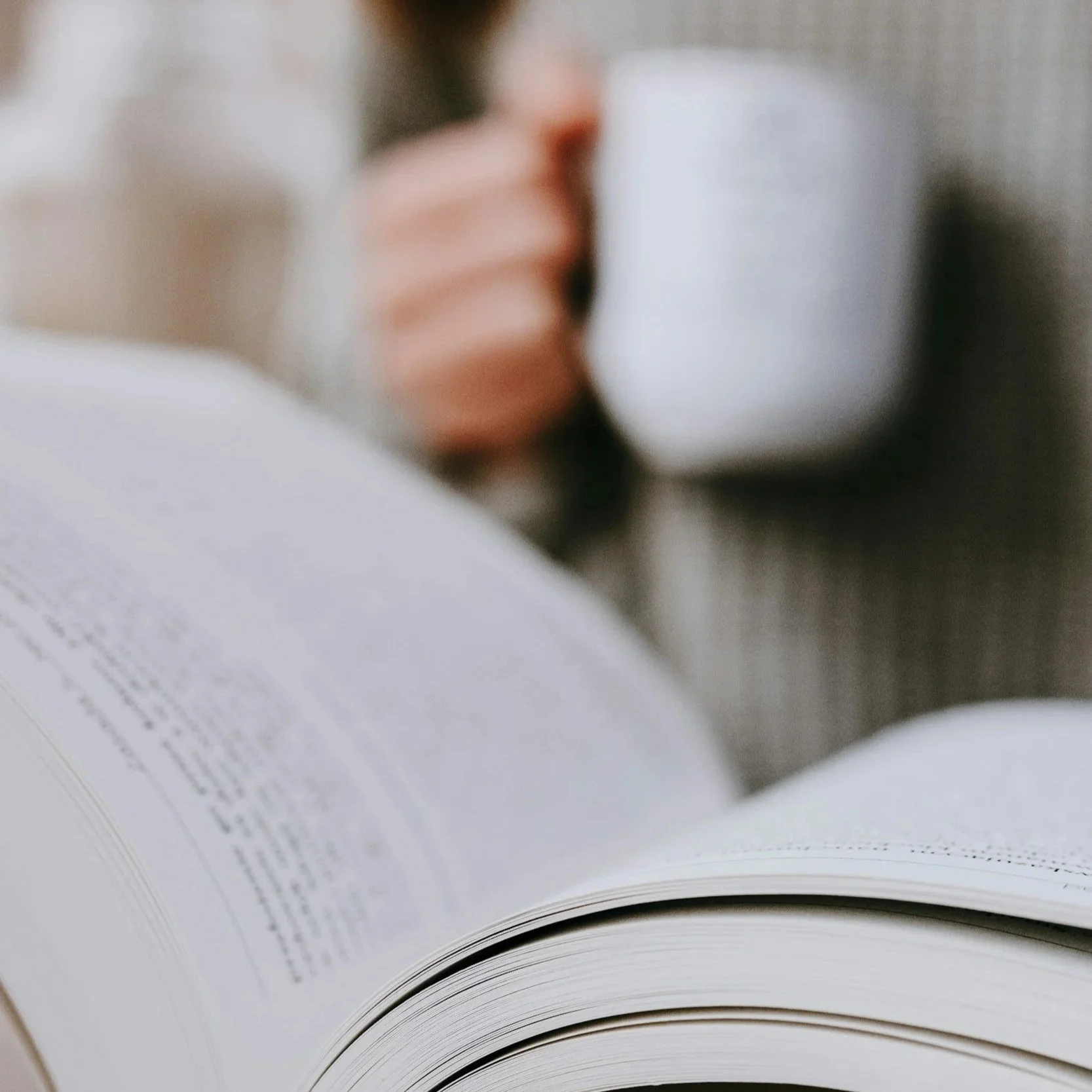 Person holding a white mug while reading a book.