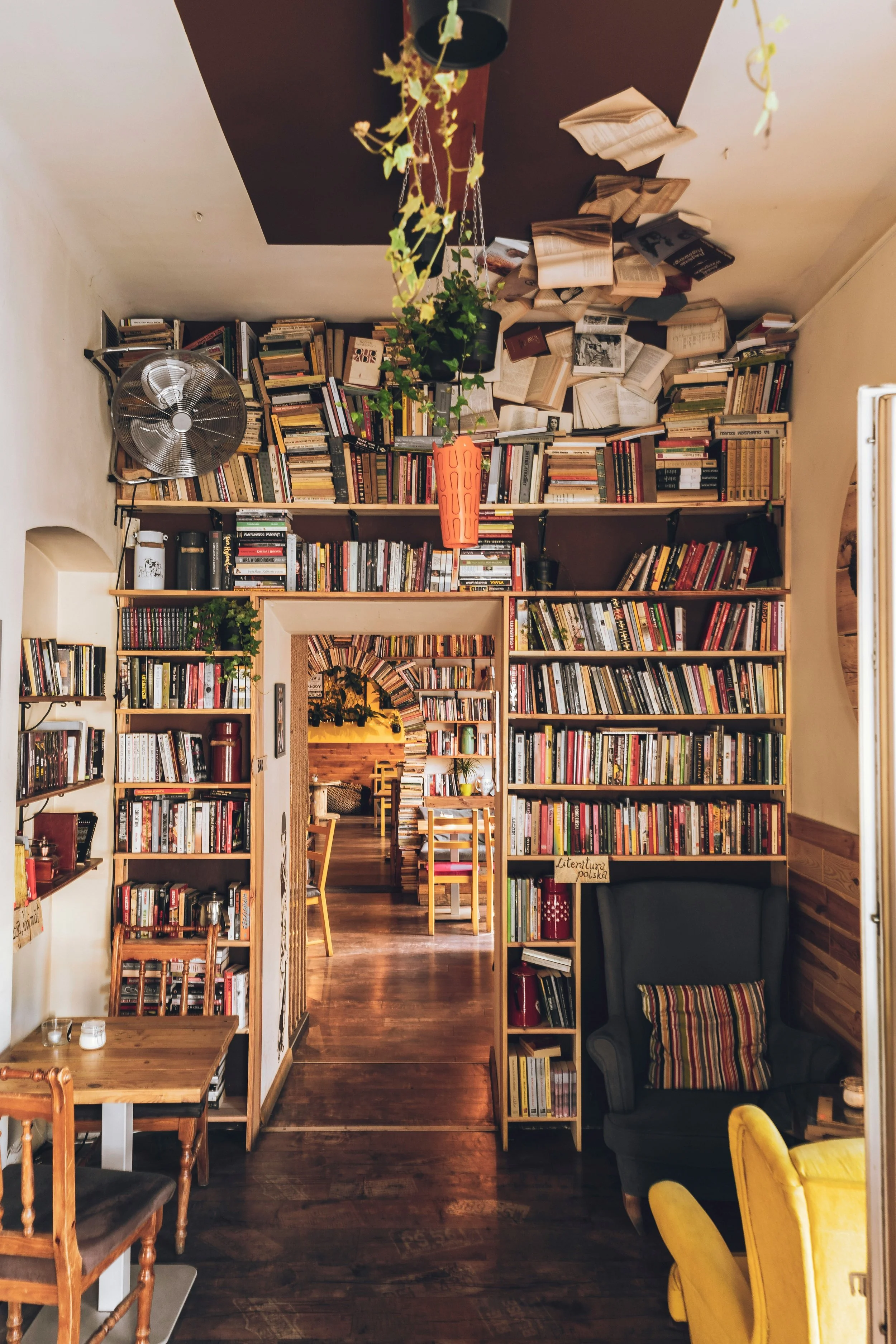 Interior of a cozy bookstore or library with bookshelves filled with books, a small table with candles, a black armchair with a colorful striped pillow, wooden chairs, and a doorway leading to another room. Plants and decor are also visible.