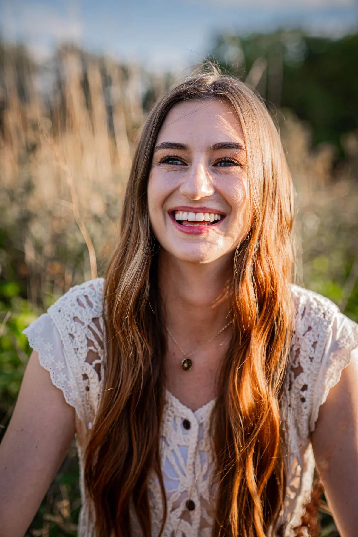 Dr Cloie Parfitt. Young woman with long red hair smiling outdoors in a field, wearing a white lace top and a necklace.