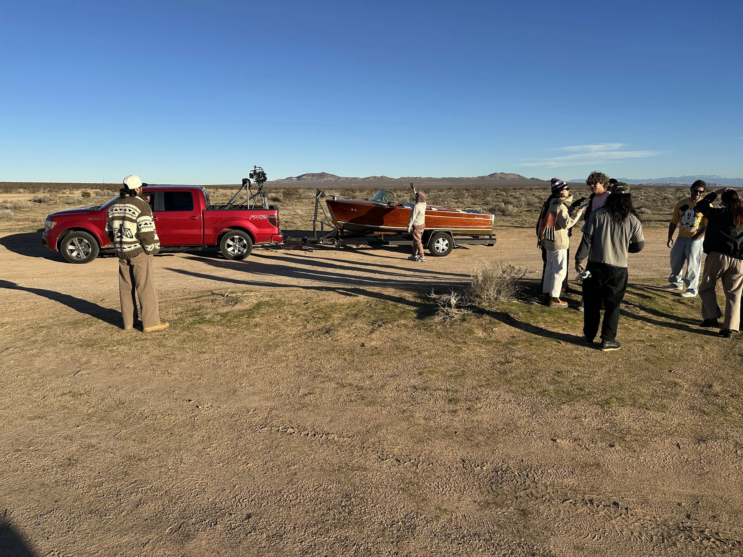 Group of people near a red truck with a boat on a trailer in a desert landscape with mountains in the background.