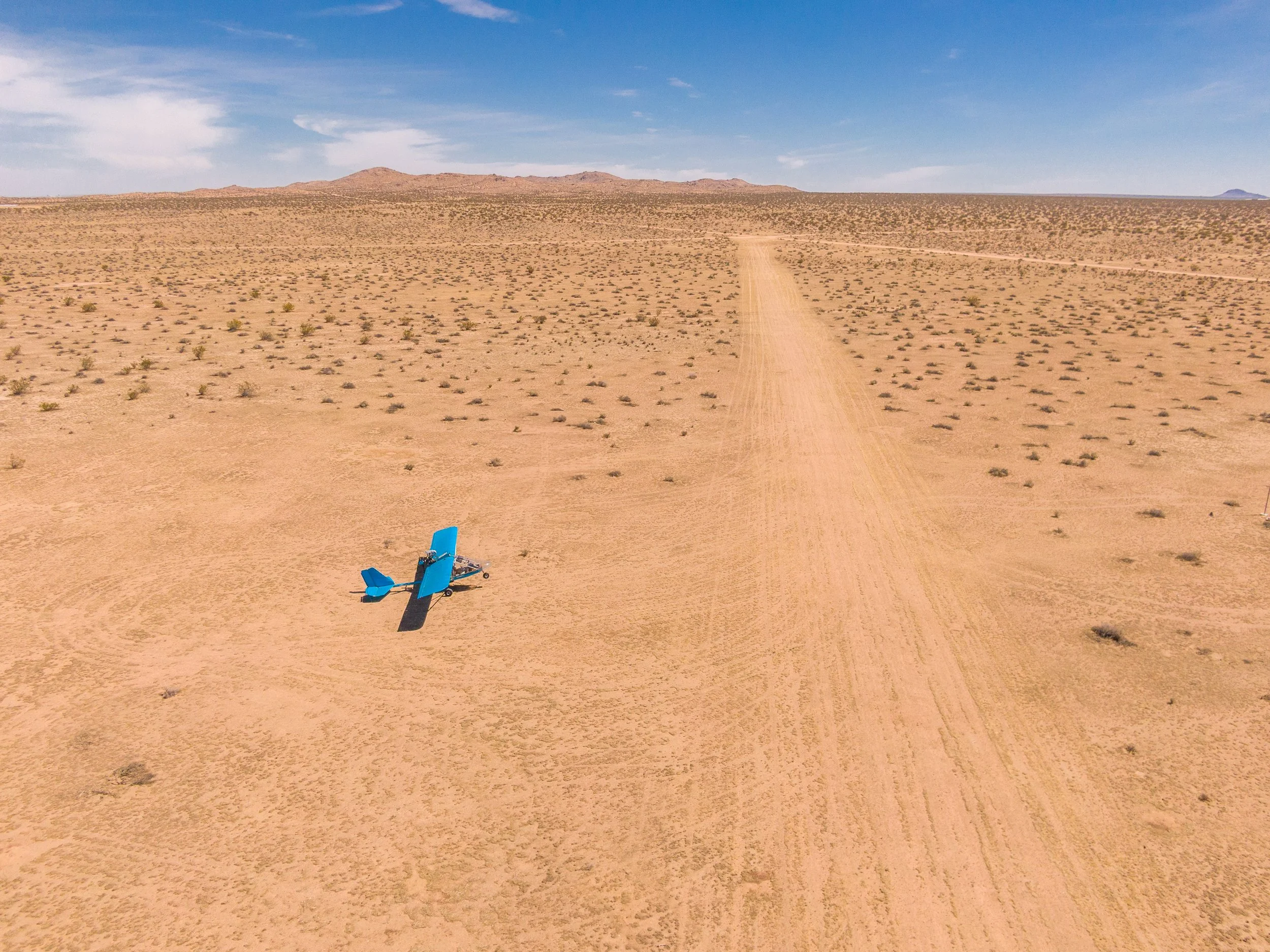 A small blue airplane crashed on a dirt runway in a desert landscape with sparse vegetation and distant mountains under a partly cloudy sky.