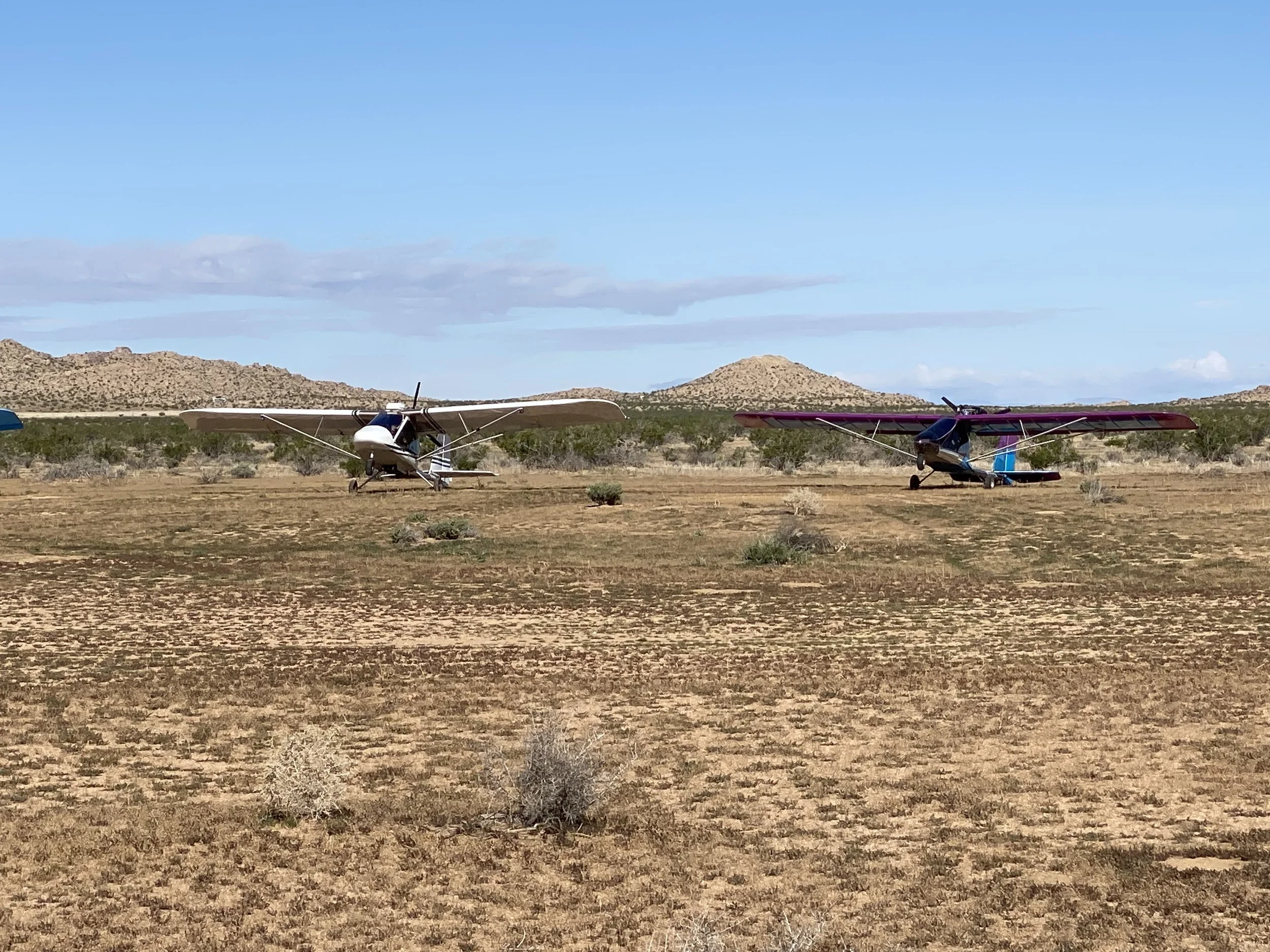 Two small airplanes parked on a dry, desert-like terrain with sparse vegetation, against a backdrop of low hills under a partly cloudy sky.