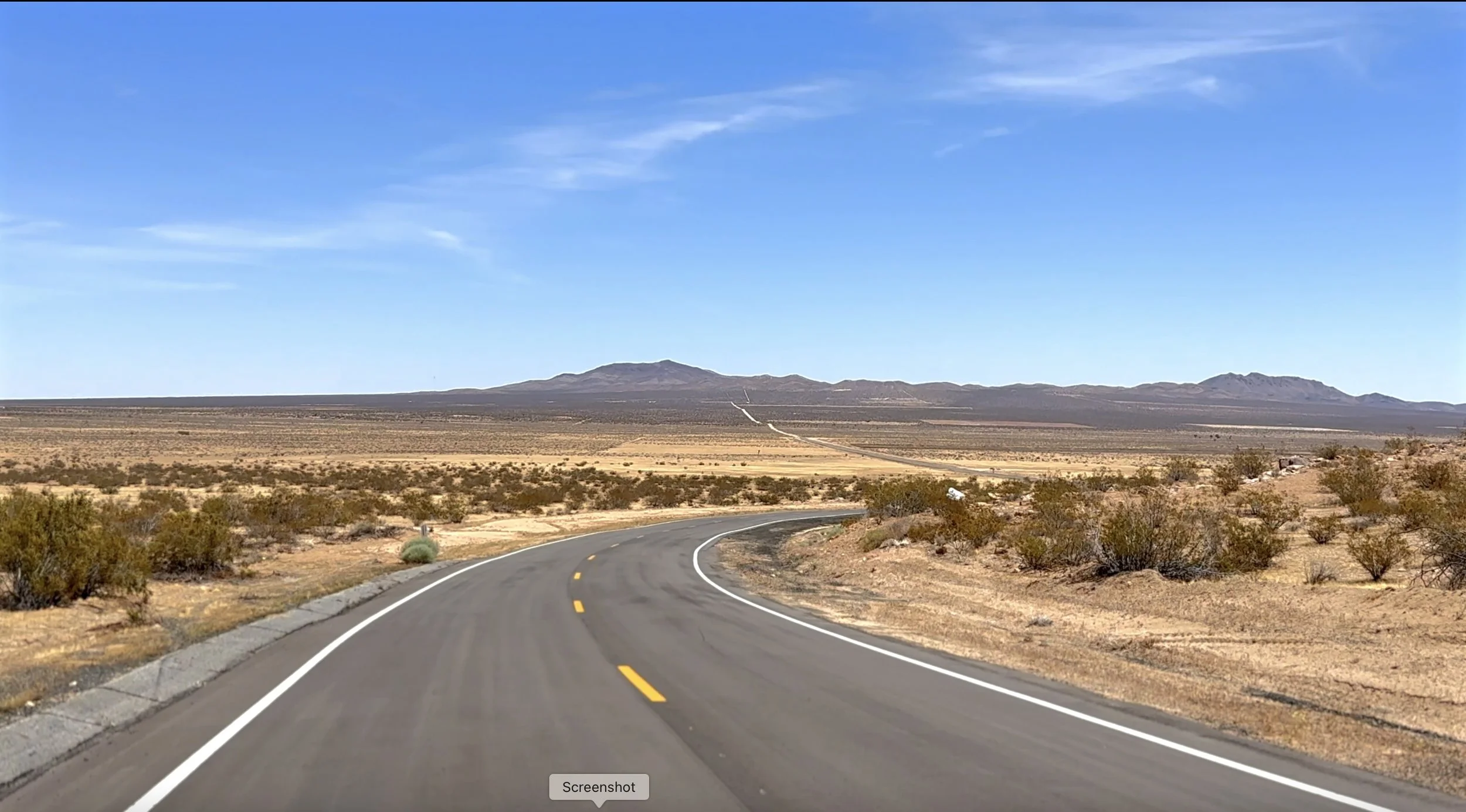Winding road through a desert landscape with sparse bushes, distant mountains, and a partly cloudy blue sky.