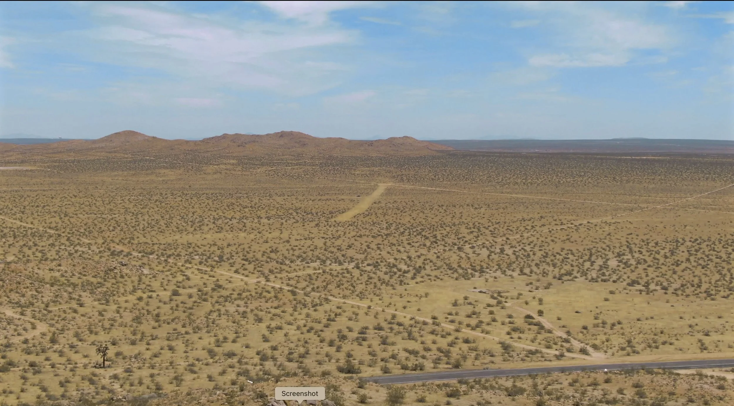 A vast desert landscape with sparse vegetation, rolling hills in the background, and a clear blue sky with scattered clouds.