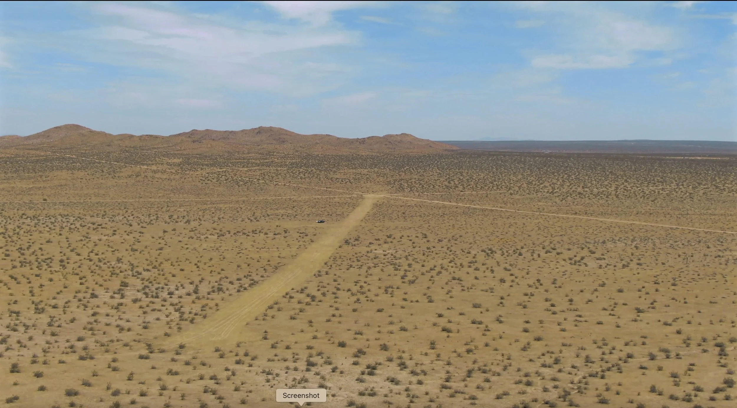 A vast desert landscape with sparse vegetation, distant mountains, a dirt road leading towards the mountains, and a vehicle on the road under a partly cloudy sky.